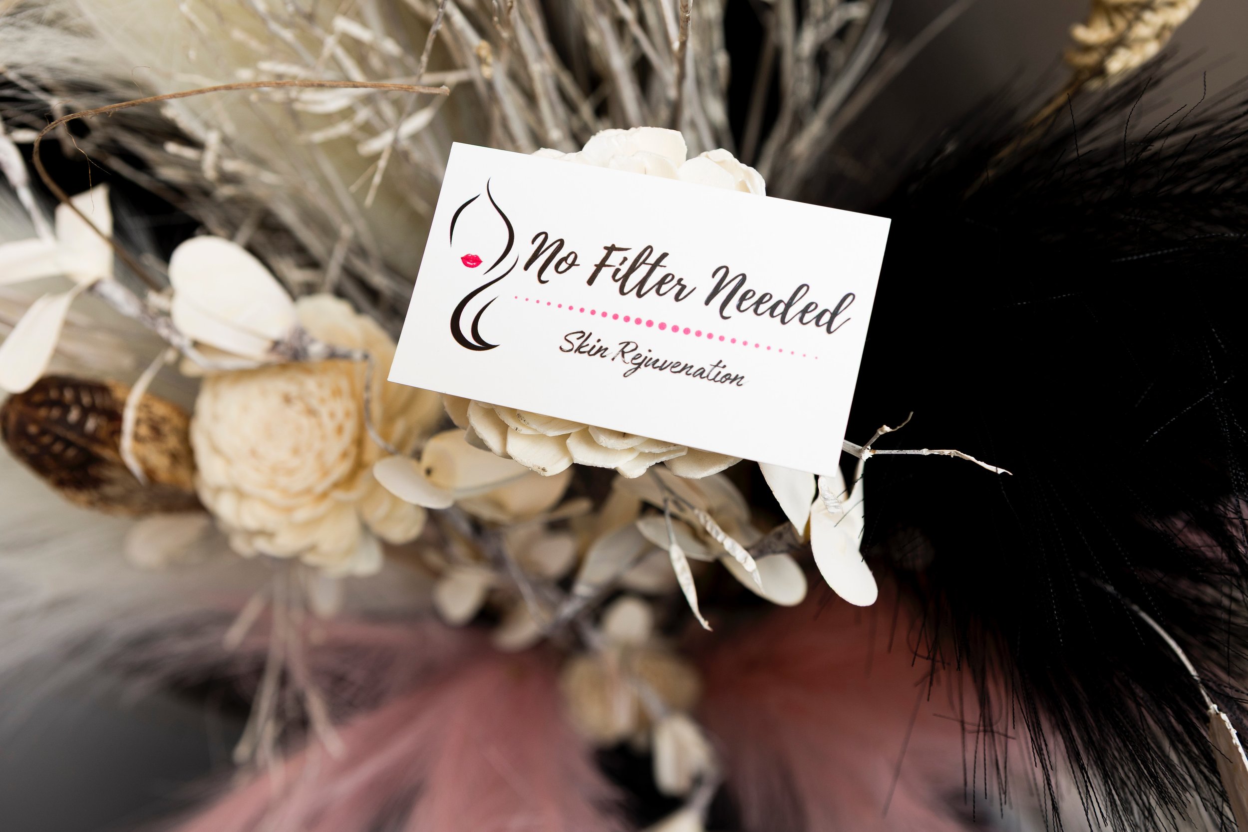 A decorative arrangement of dried flowers and feathers with a sign that reads 'No Filter Needed, Skin Rejuvenation'.