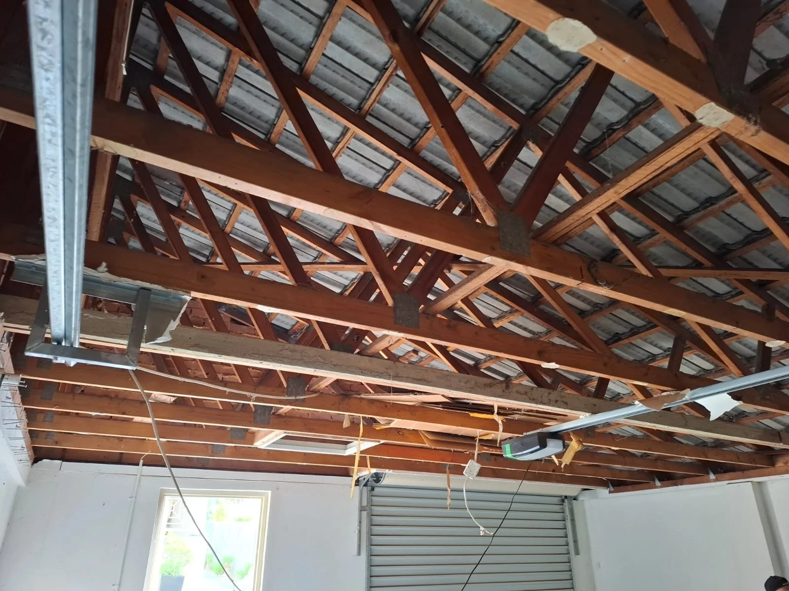 Inside of a garage with exposed wooden ceiling beams and roof trusses, a roller garage door, a window, and construction wiring.