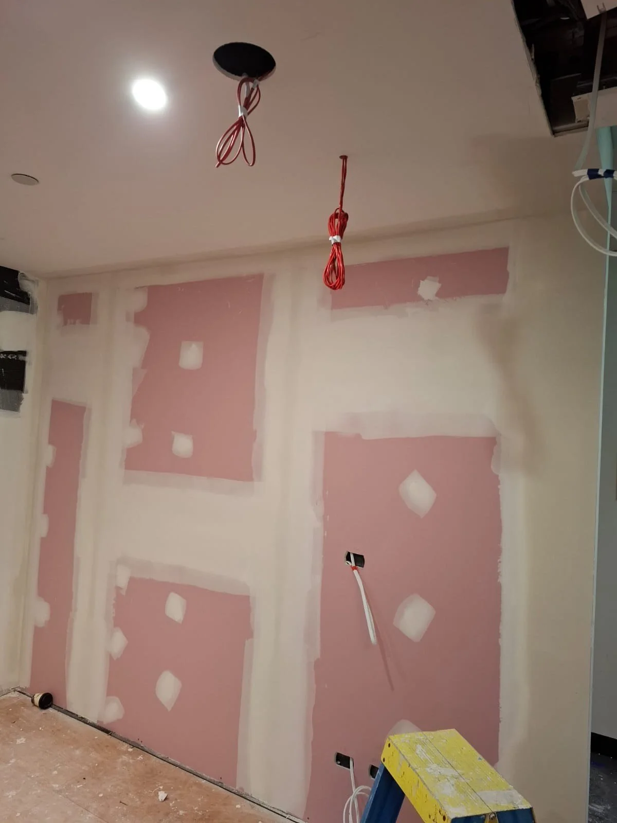 Interior wall under construction with pink drywall panels and drywall joint compound, electrical wiring hanging from the ceiling, and construction tools visible.