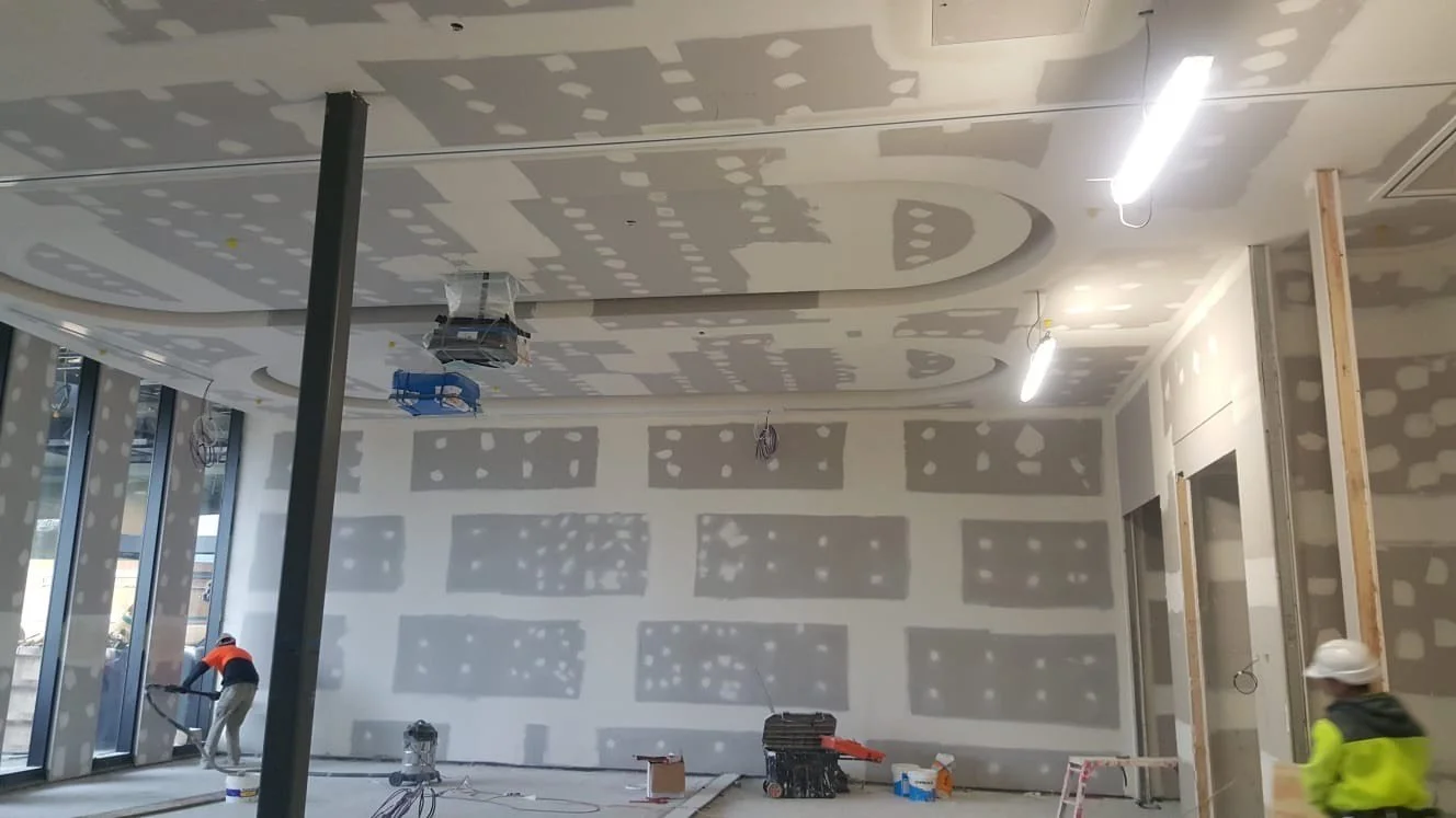 Interior of a building under construction with workers and light fixtures, drywall with joint compound in progress.