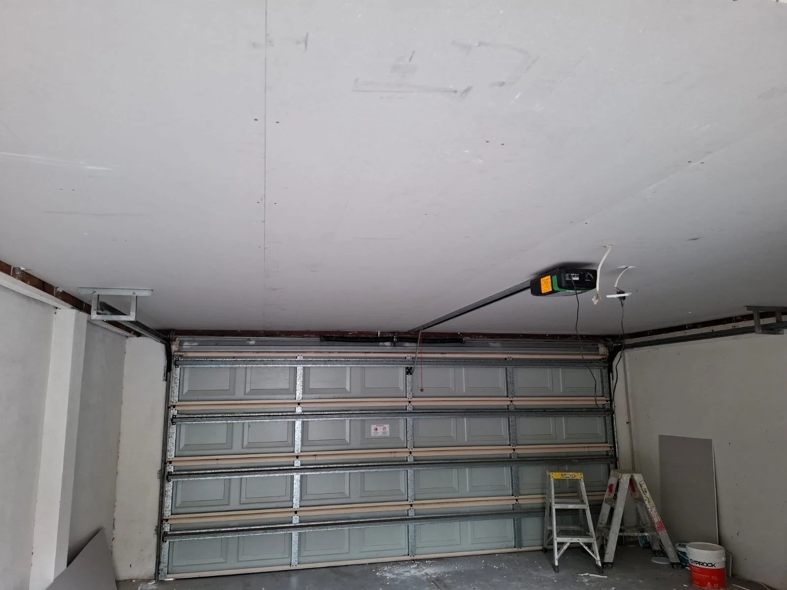 A garage with a closed metal garage door, an overhead garage door opener, two step ladders, a bucket, and construction supplies.