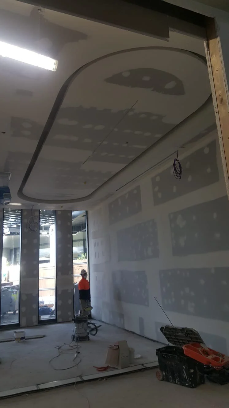 Interior of a building under construction with drywall installation, a drywall finisher in an orange and dark jacket, and construction tools on the floor.