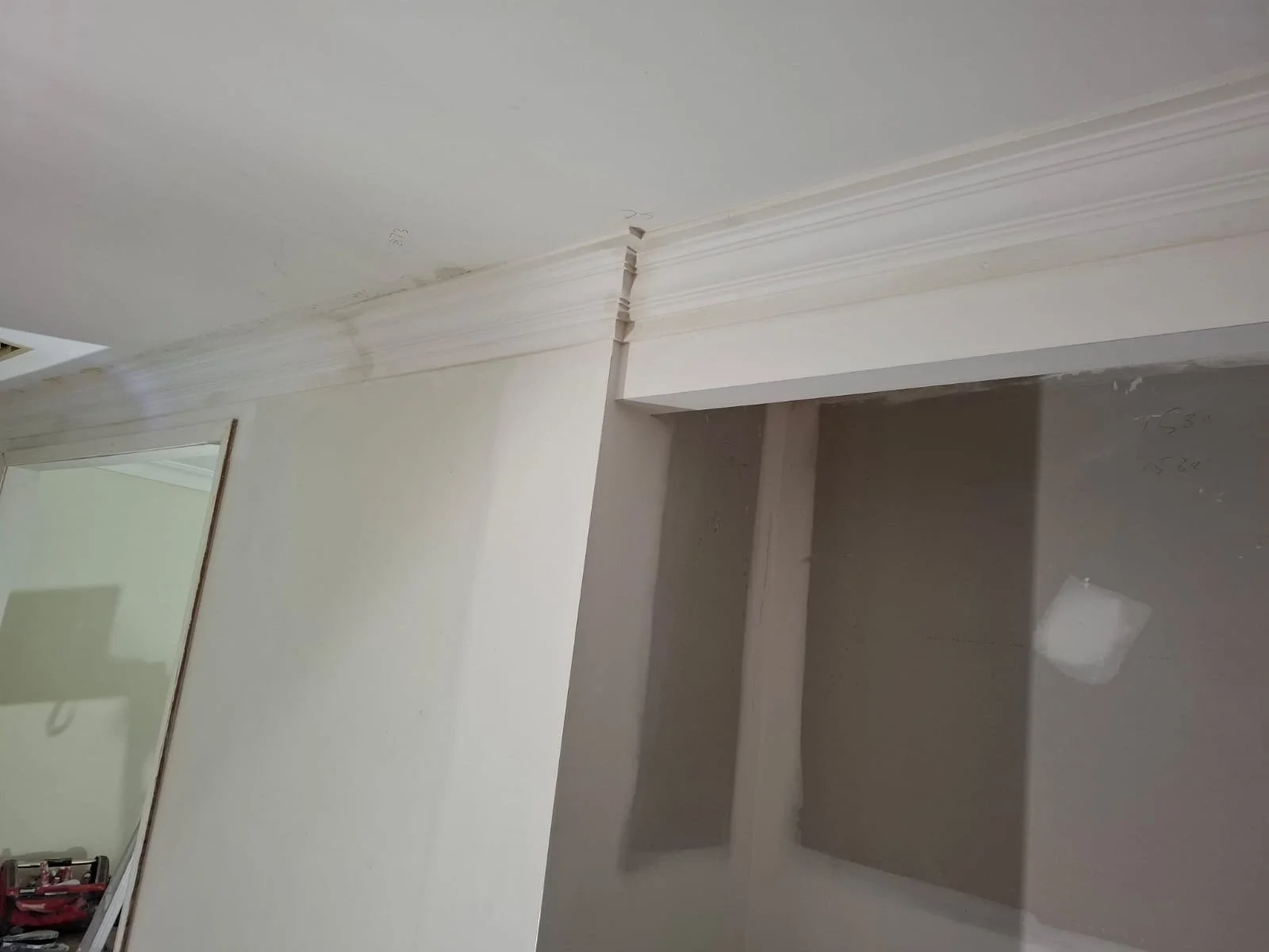 Interior of a room under construction or renovation, showing a corner with drywall walls, crown molding, and some tools and materials in the background.