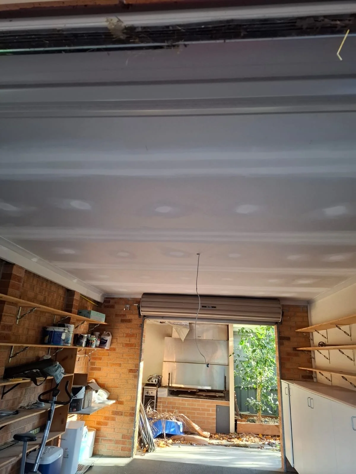 Indoor garage with shelves on the left holding various containers, for example, paint, and tools. A white portable refrigerator is on the floor. In the background, an open garage door reveals greenery outside and a garden bed with a small tree. The c