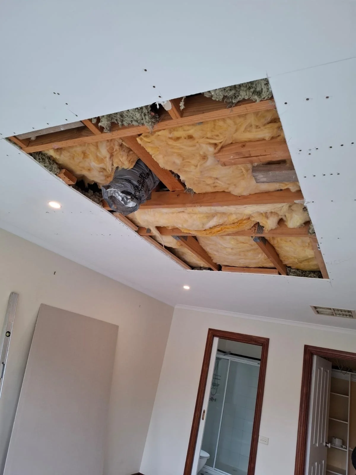 Ceiling with a large rectangular section removed, exposing wood framing, yellow insulation material, and ductwork, in a room with beige walls and adjacent bathroom.