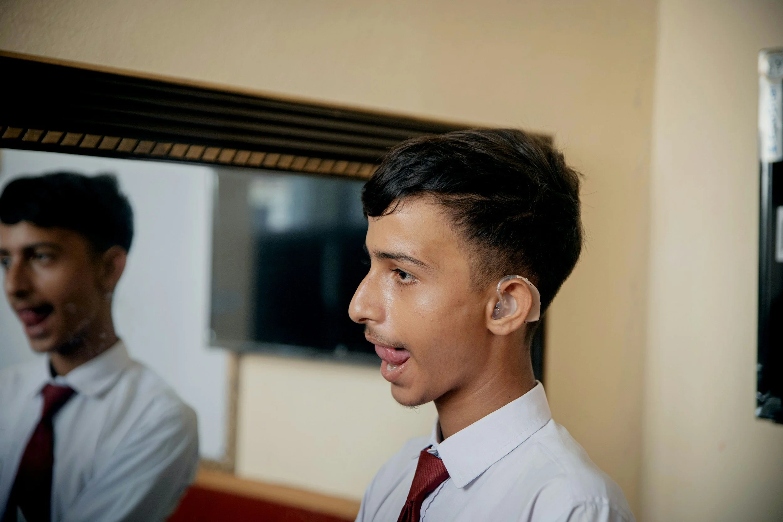 Two young men in school uniforms with white shirts and red ties, standing indoors, one with an ear prosthetic and the other smiling, with a mirror and television in the background.