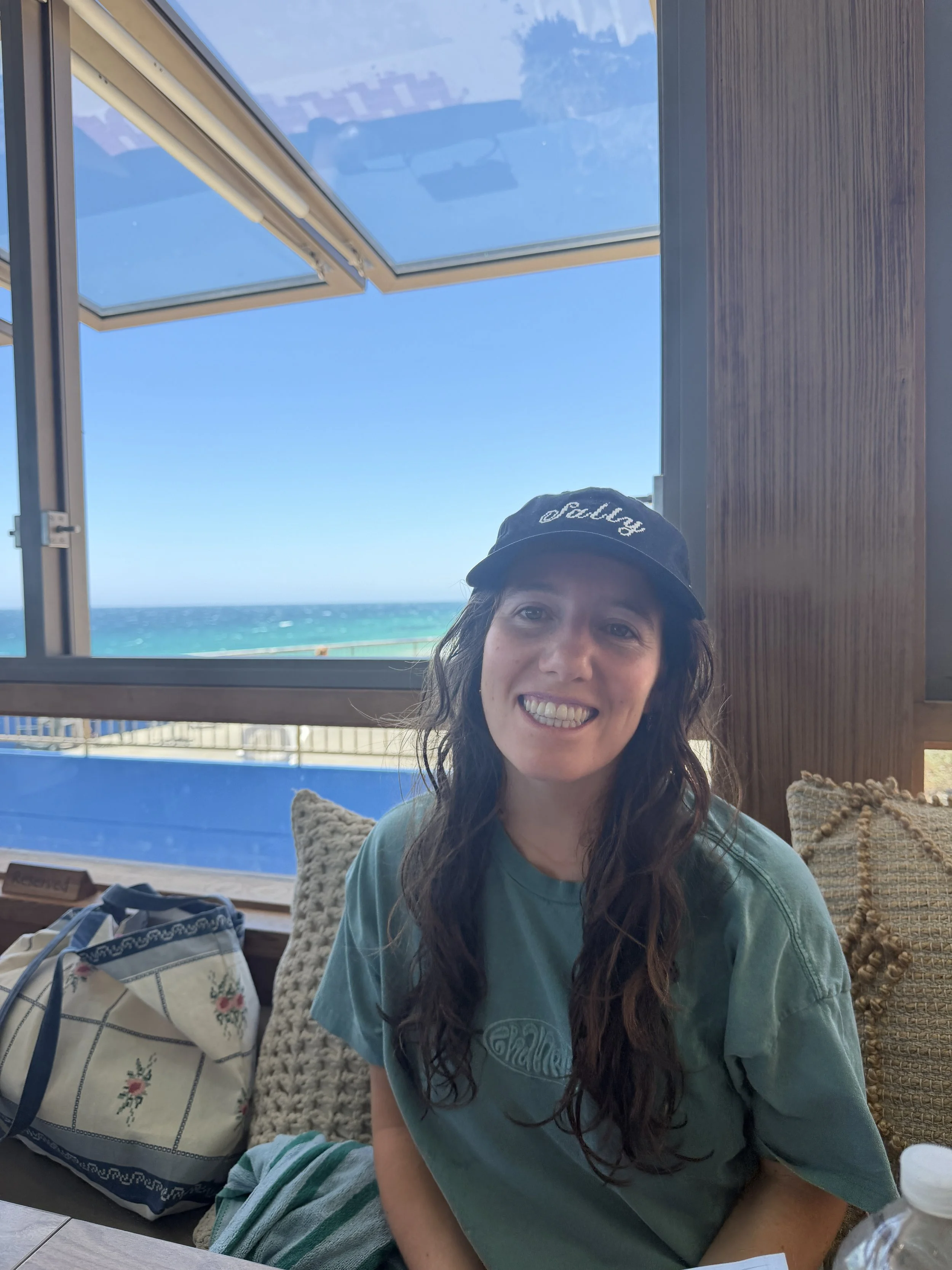 A woman with long curly hair wearing a black cap and green t-shirt, smiling while sitting inside a building near a large open window with an ocean view.