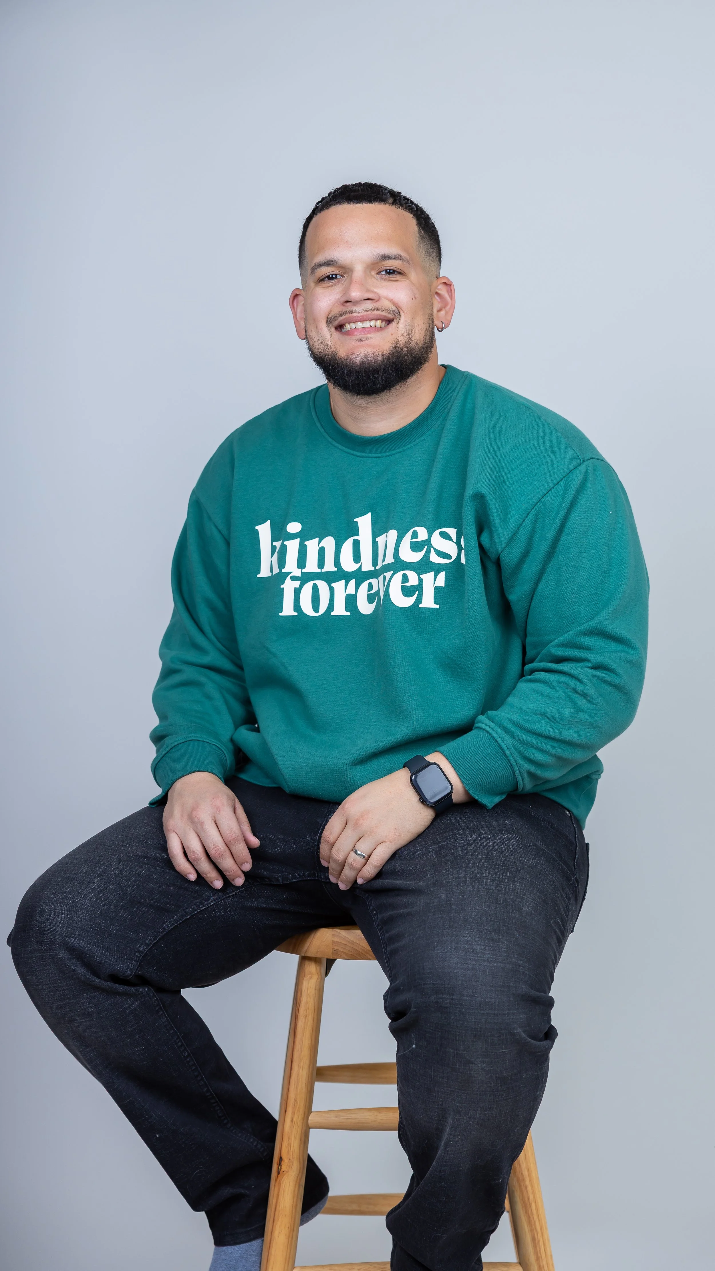 A man with short dark hair, a beard, and earrings smiling, sitting on a wooden stool against a light grey background. He is wearing a teal sweatshirt with white text reading 'kindness forever,' black jeans, a smartwatch, and a wedding band.