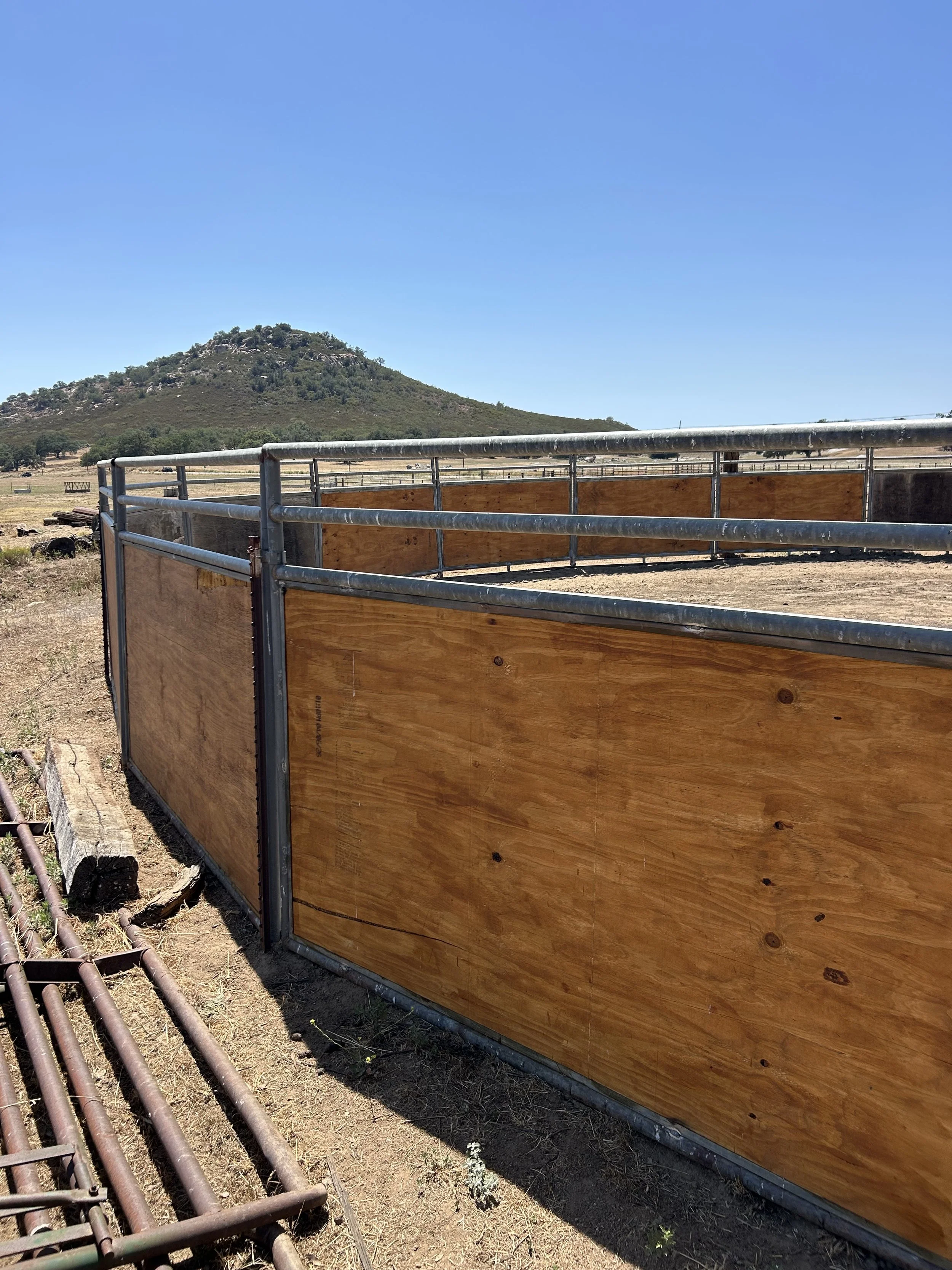 A metal and wood cattle pen on dry ground with a hill covered in vegetation in the background under a clear blue sky.