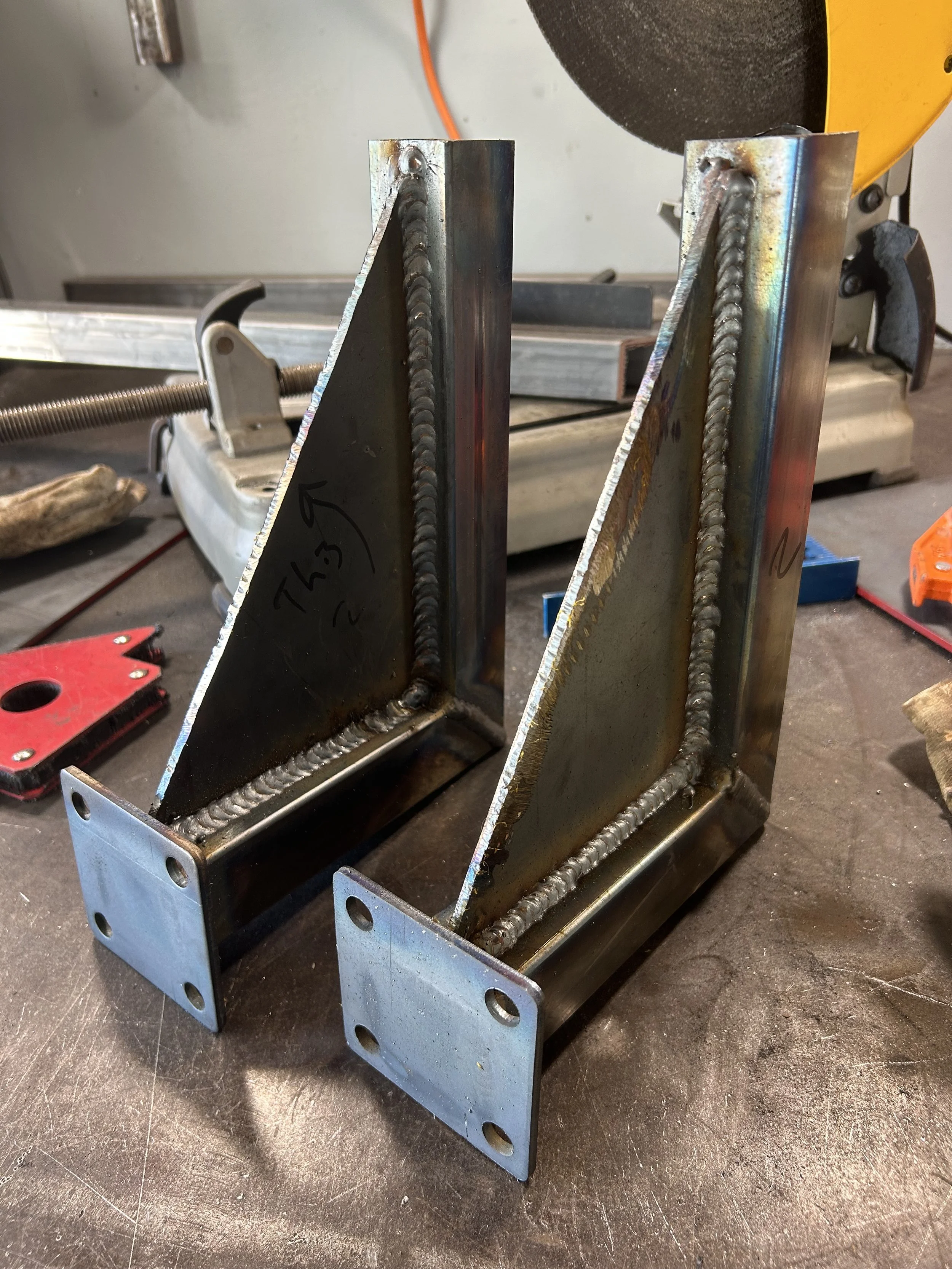 Metal components welded together to form a structure, with visible welds on the steel plates. In the background, a yellow grinder and various tools are on a workbench, indicating a workshop setting.