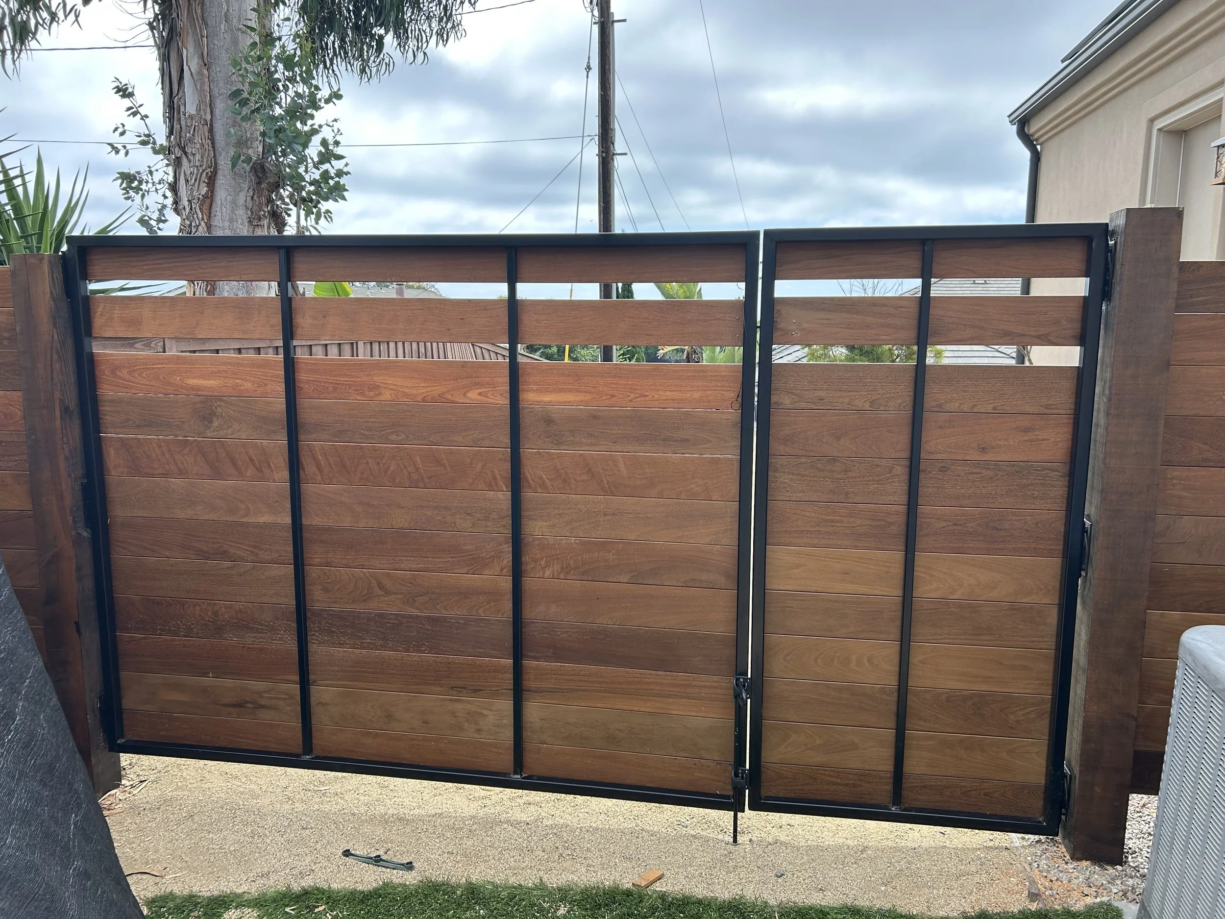 A newly installed wooden fence with black metal framing. There's a large tree with a textured trunk and lush green leaves to the left, and a house with a light-colored exterior on the right. Several power lines run across the cloudy sky, and the grou