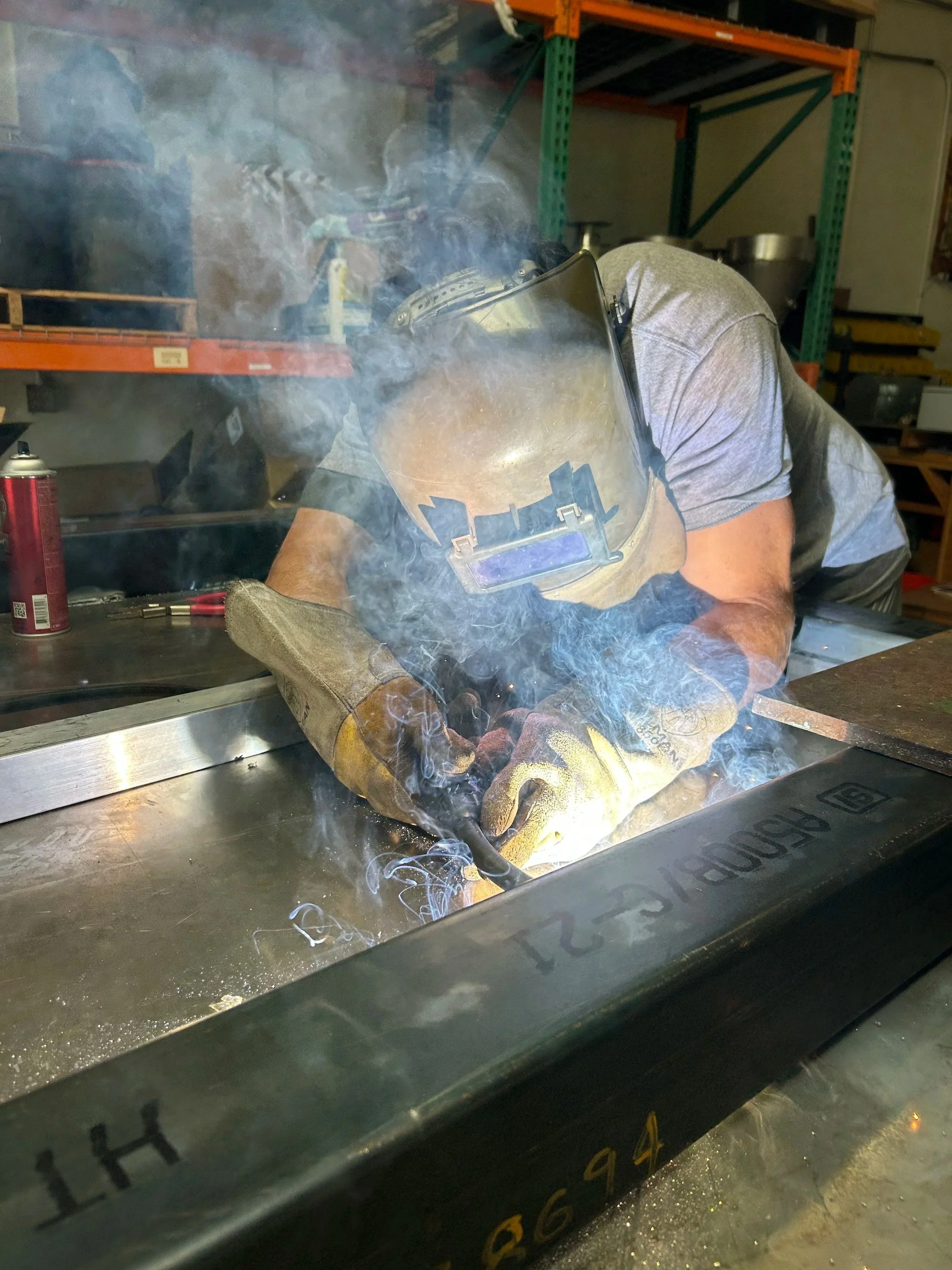 A person welding at a workshop workspace, wearing a welding helmet and gloves, with smoke and sparks visible around the welding area.