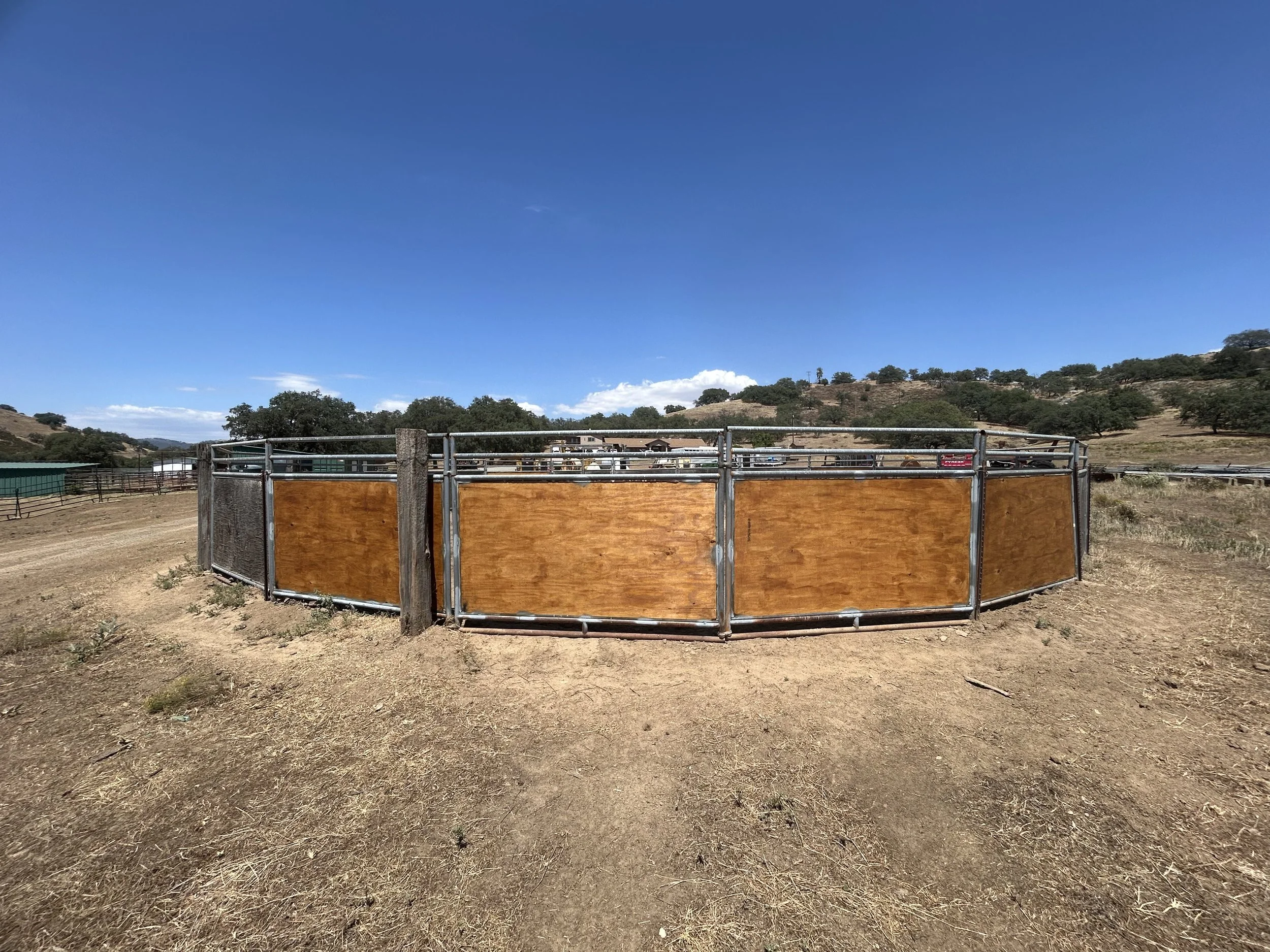 A fenced outdoor area with dry brown grass and hills in the background under a clear blue sky.