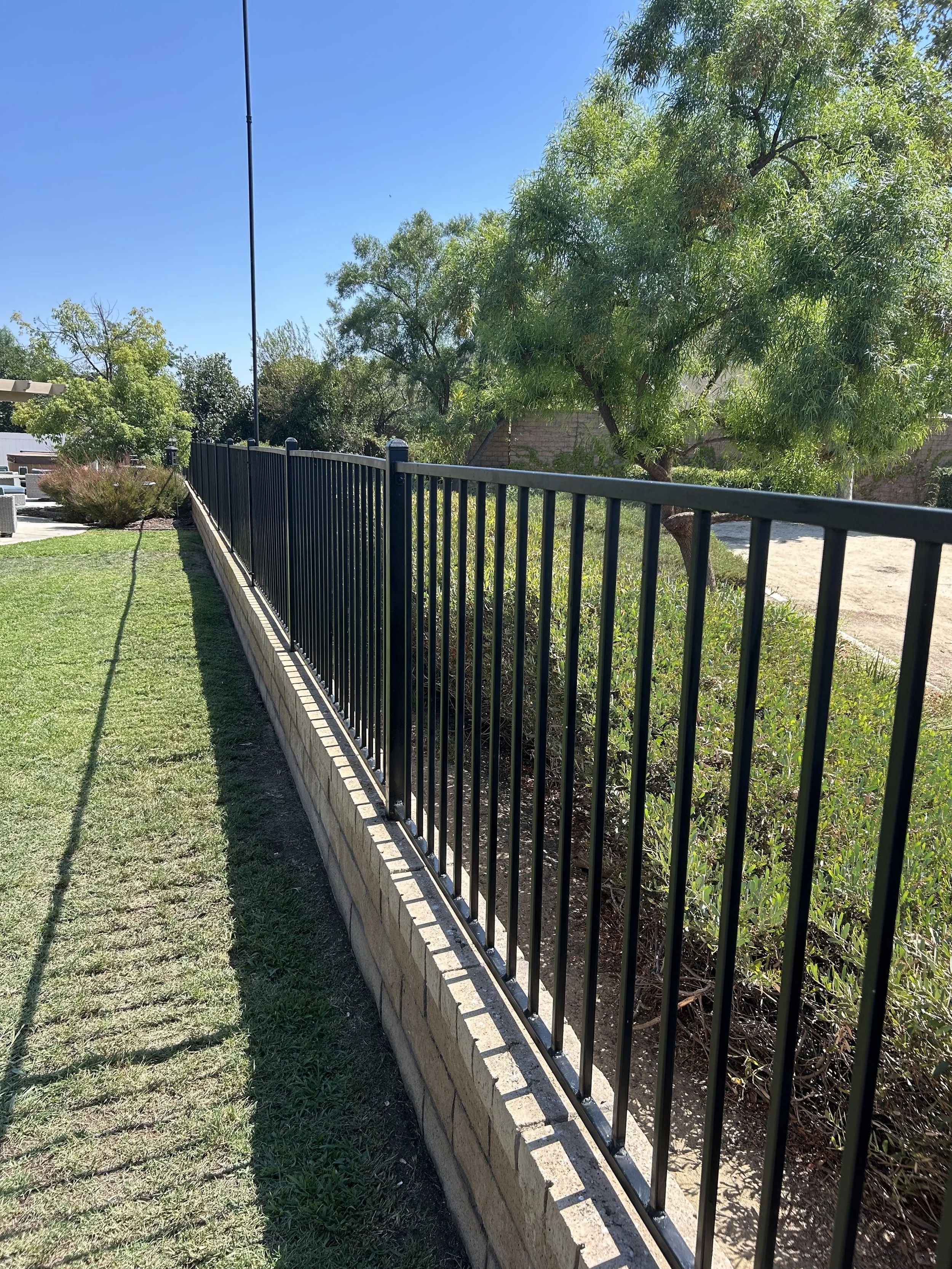 A black metal fence runs along a concrete base, bordering a grassy area and a garden with bushes and trees. The sky is clear and blue.