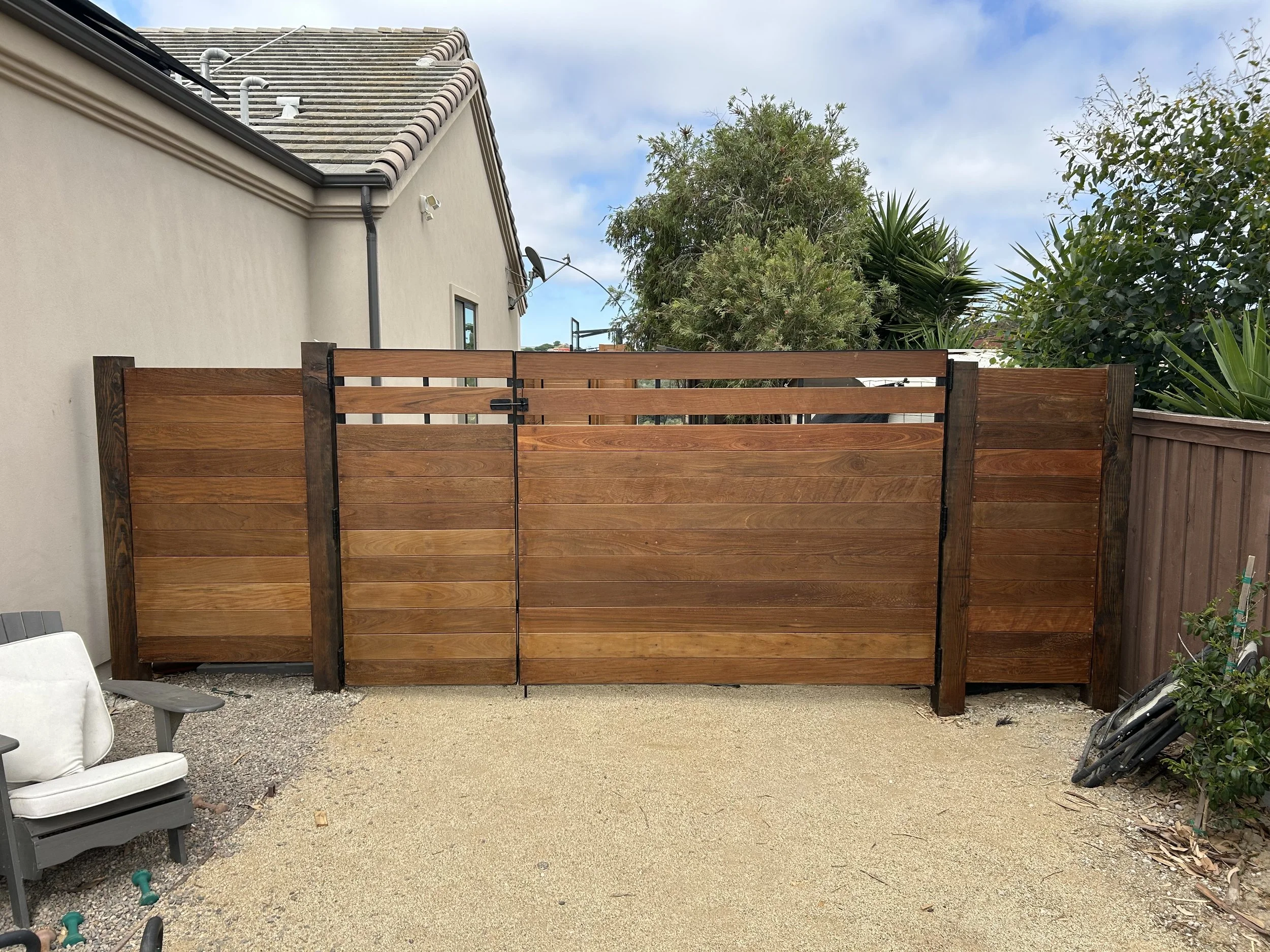 Backyard with a wooden fence, a beige house wall, trees, and a gravel ground with some outdoor furniture.