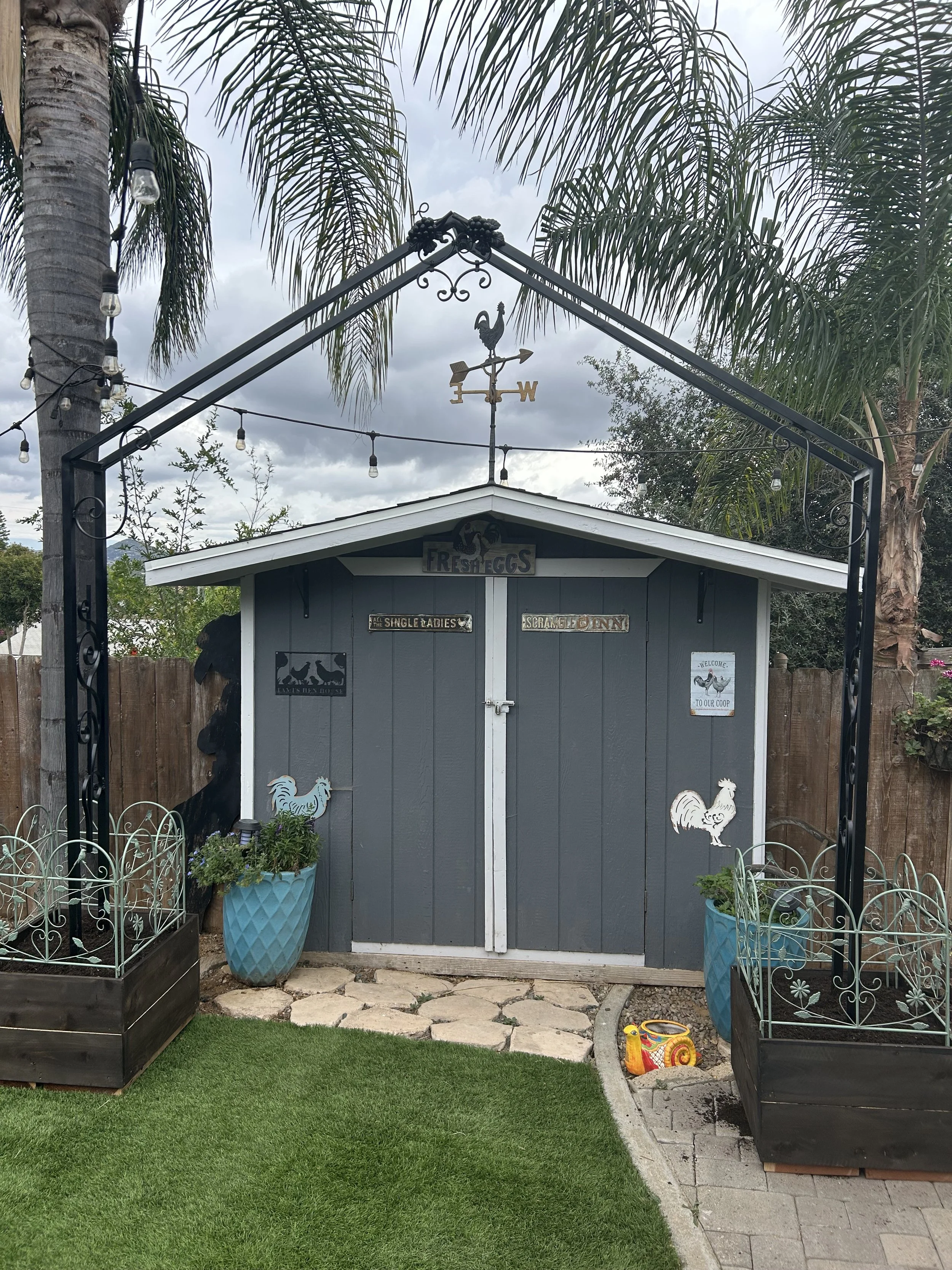 A small gray garden shed with decorative signs and rooster ornaments, positioned behind a black metal archway with a weather vane featuring a rooster, and surrounded by planters and a manicured lawn.