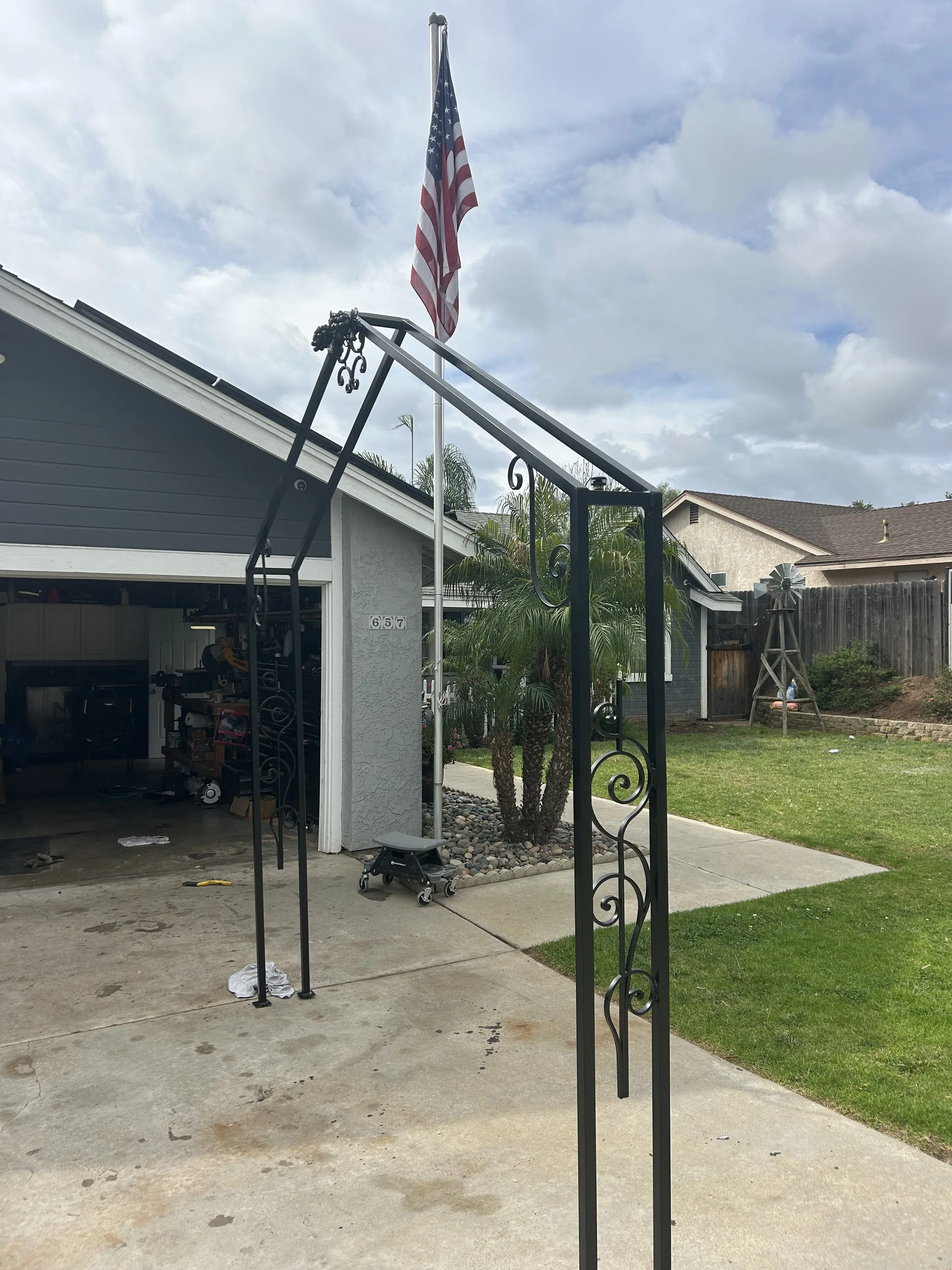 A backyard with a flagpole and an American flag; a house with gray siding and white trim; a black wrought-iron gate frame in the foreground; a small palm tree and a garden bed with rocks; a wooden windmill decoration; cloudy sky.