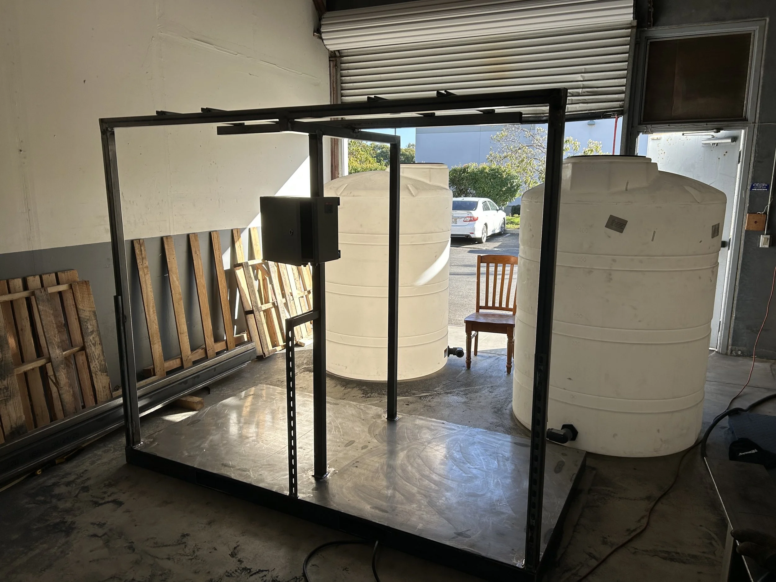 Inside a garage with two large white water tanks, wooden pallets, and a metal table frame. A chair and a small black electrical box are also visible.