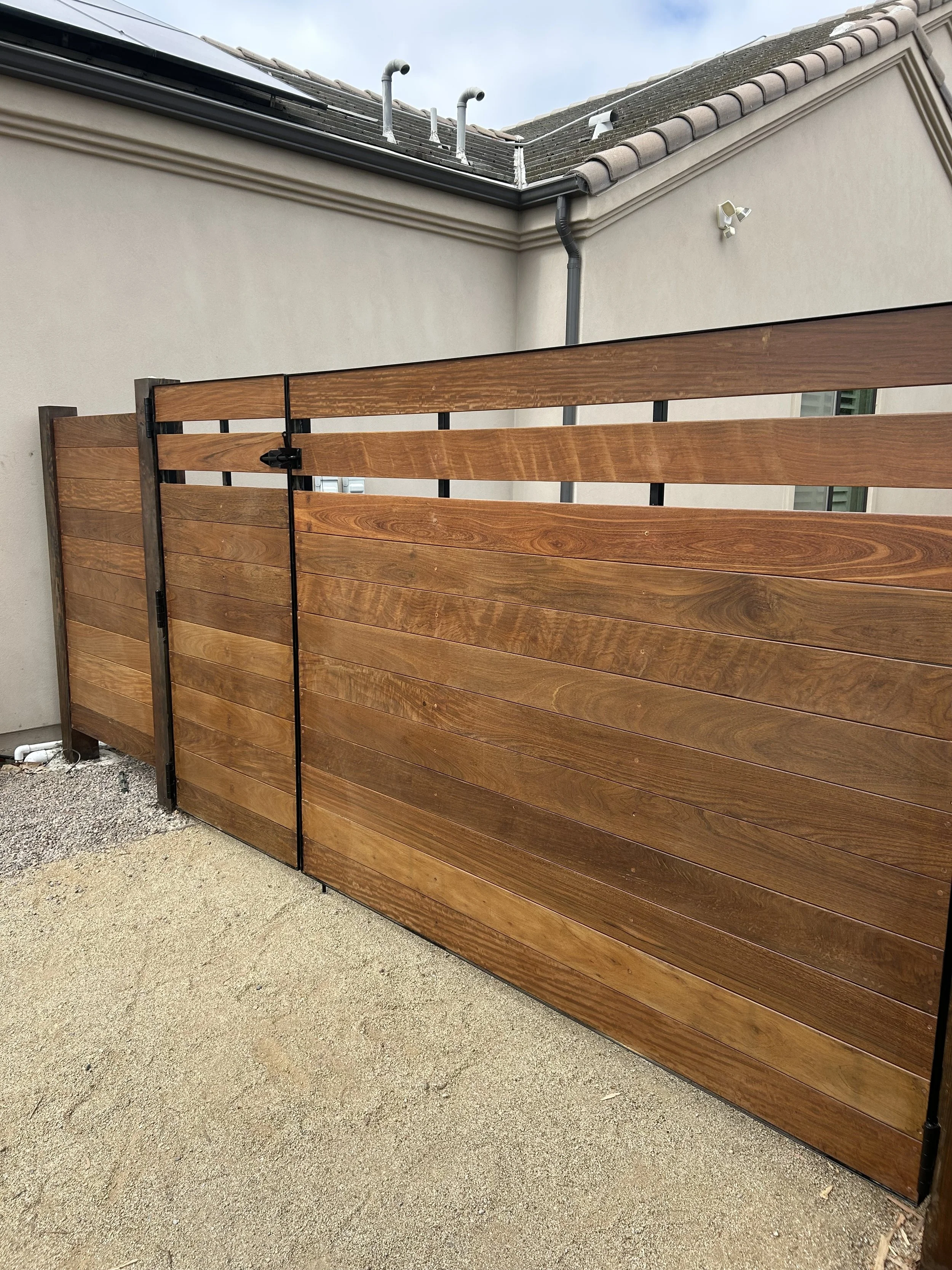 A wooden fence with horizontal planks and a black metal latch in front of a beige house wall. In the background, part of a tiled roof with two metal vents, a security camera, and a gray sky are visible.
