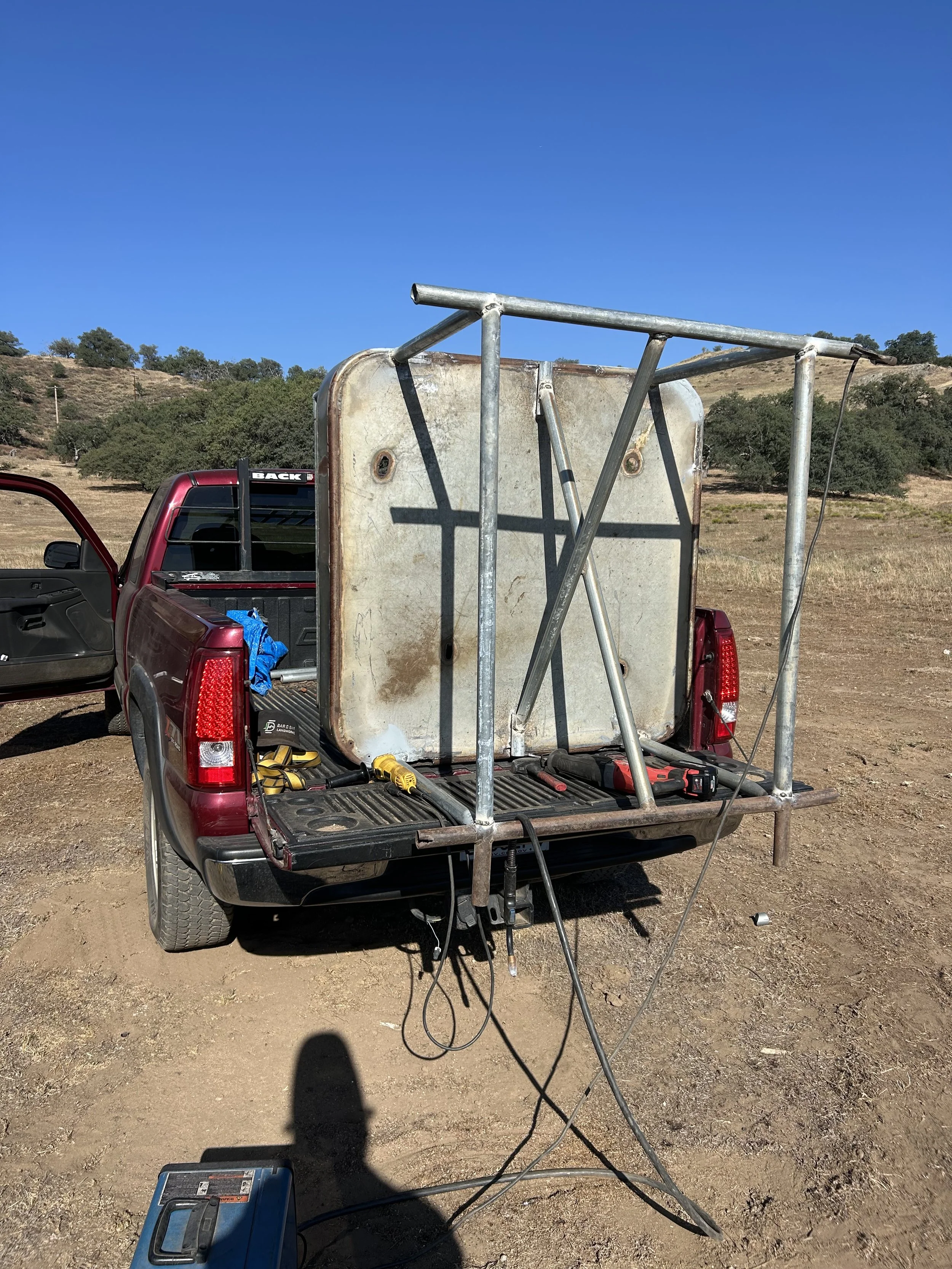 Back of a red pickup truck with its tailgate down, holding a large metal frame, a weathered rectangular metal box, and various tools including a wrench and a cordless drill. The scene is outdoors on a dirt ground with a grassy hillside and trees unde