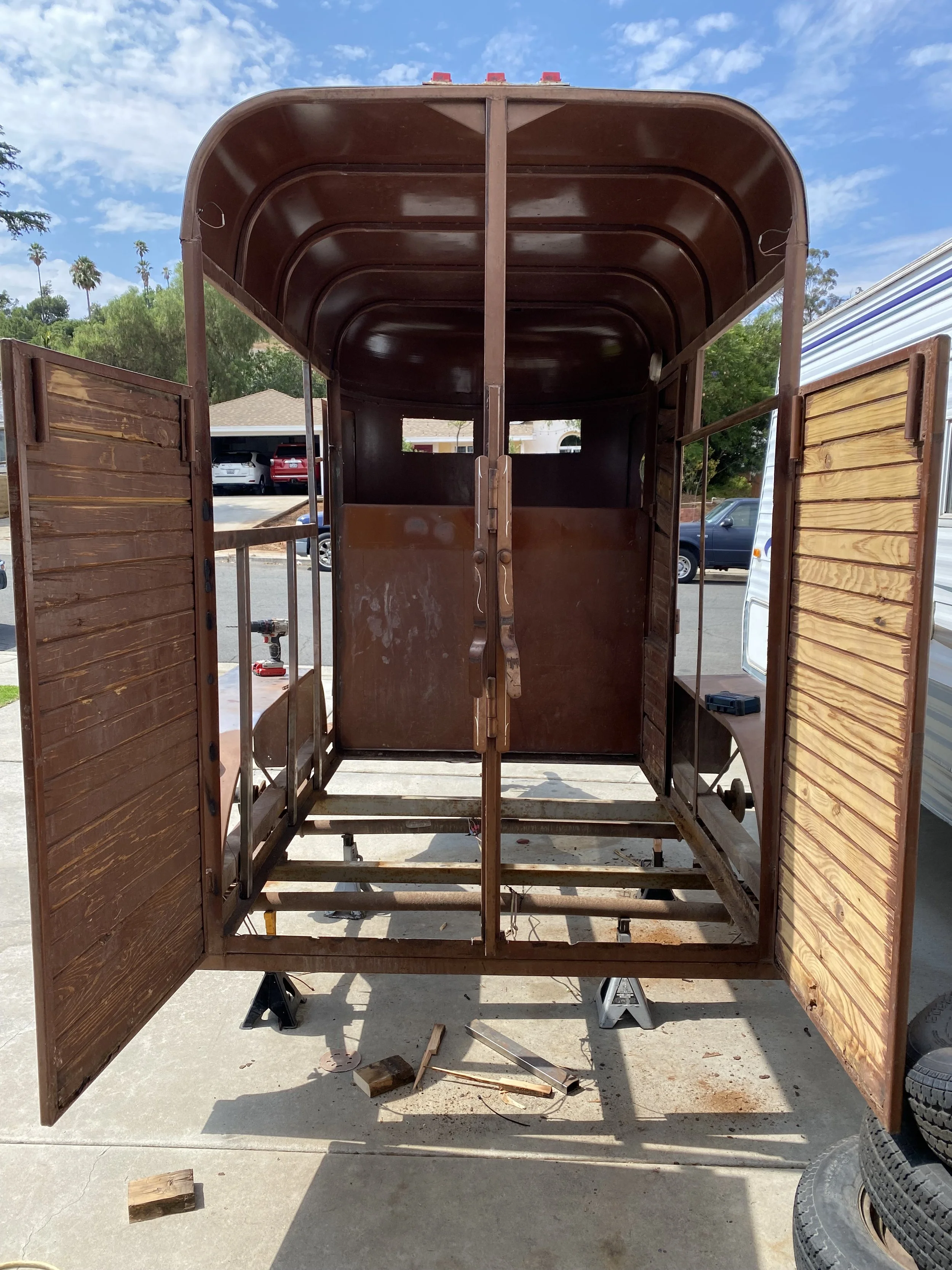A wooden trailer with open double doors, parked outside on a driveway, with tools and wood pieces underneath, and a blue sky with clouds.