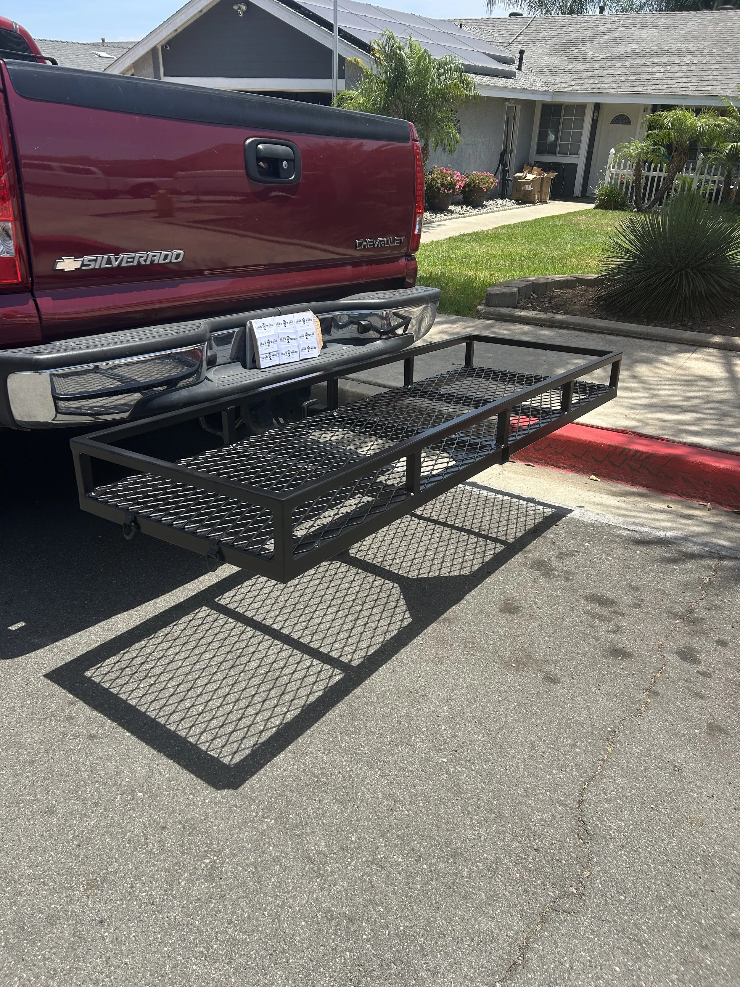 The back of a red Chevrolet Silverado pickup truck parked in a driveway with a black metal utility cart attached. The background includes a house with solar panels on the roof, palm trees, potted plants, and a small garden bed.