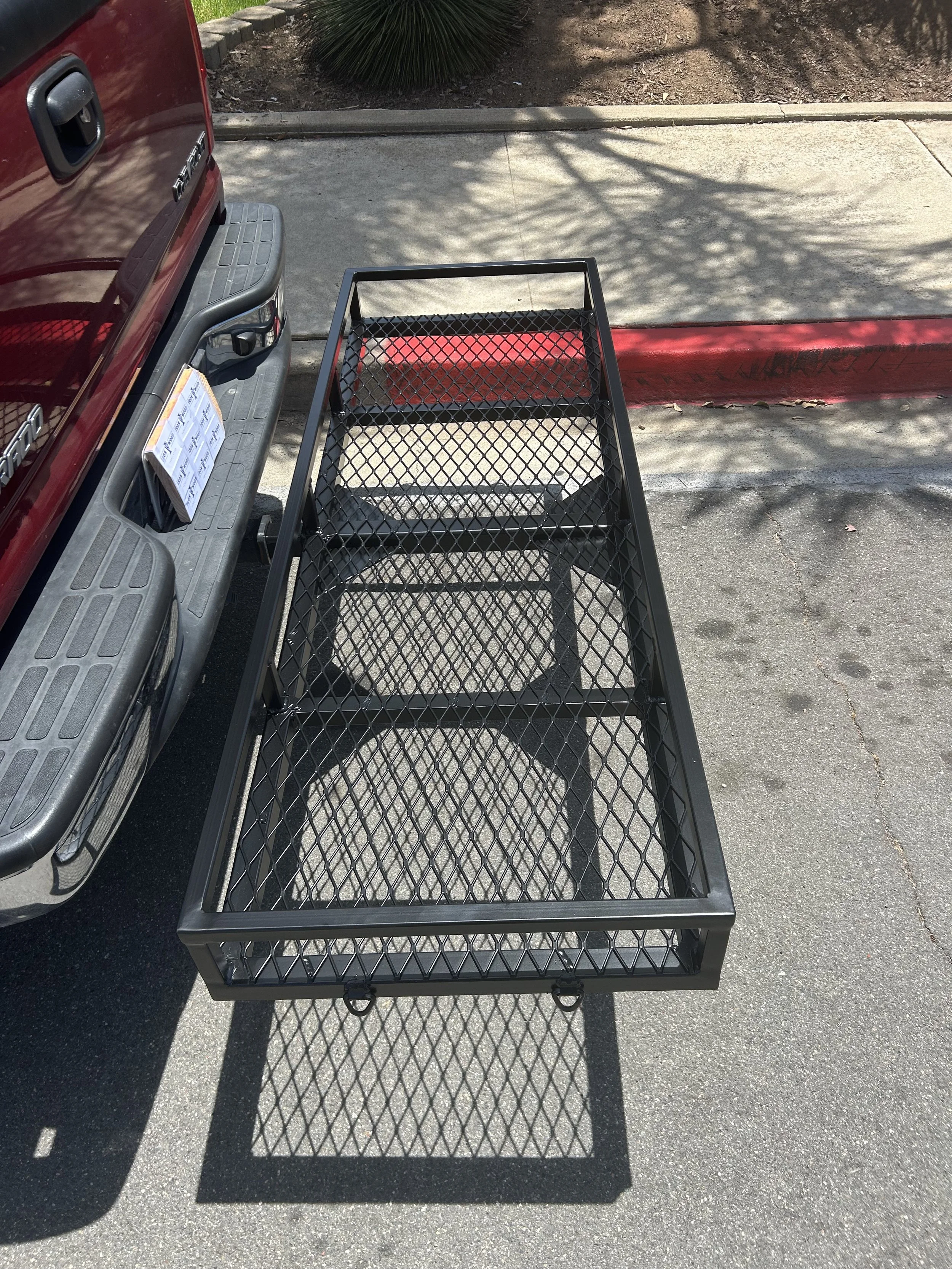 Black metal utility cart with a diamond pattern on the bottom shelf, placed on a parking lot beside a concrete curb and a red painted line, with a shadow of tree branches overhead.