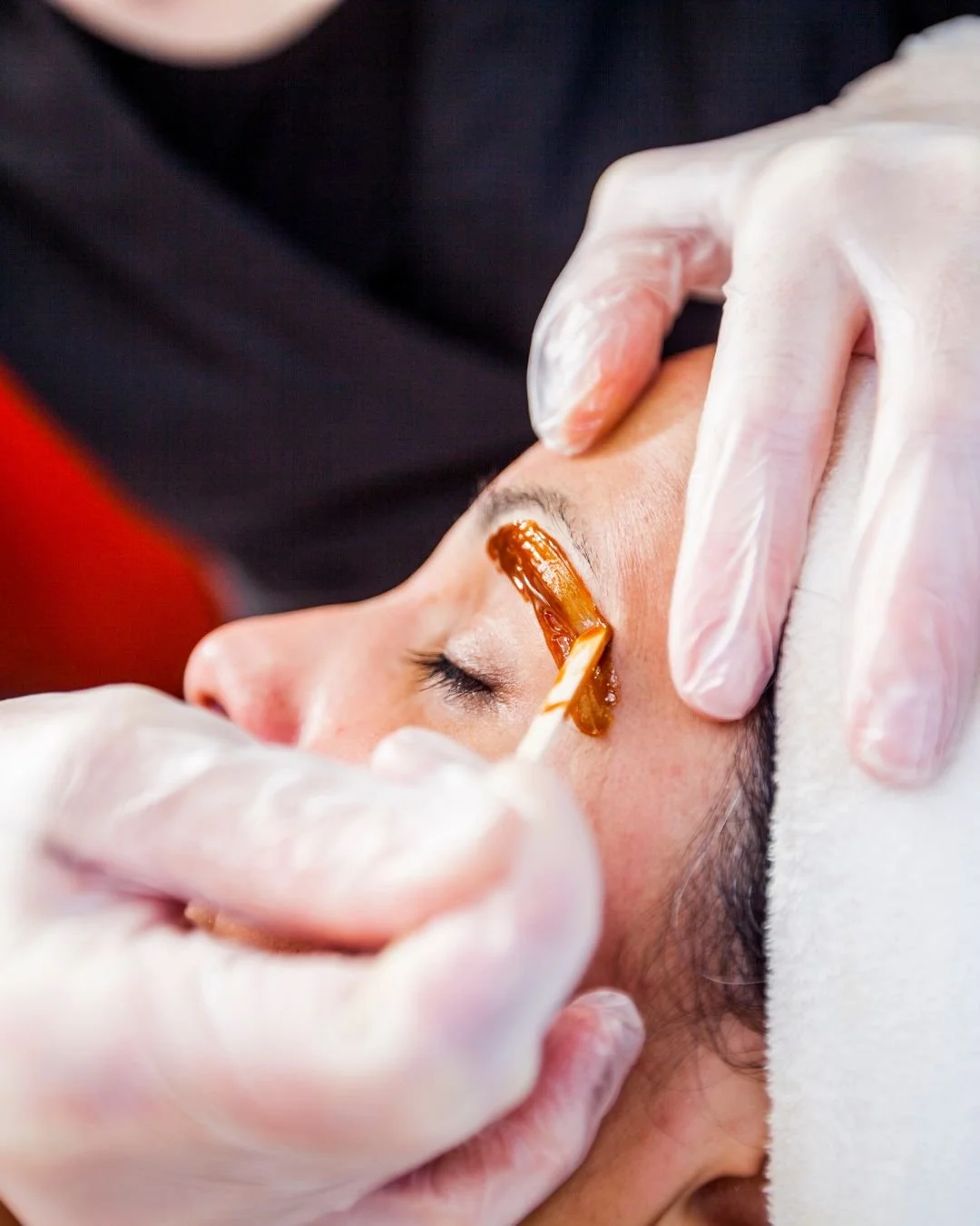Licensed aesthetician applying warm eyebrow wax to a client's brow with a wooden spatula