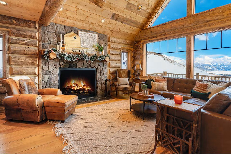 Cozy rustic living room with large windows, wooden walls, and a stone fireplace decorated with garland, overlooking snow-covered mountains.