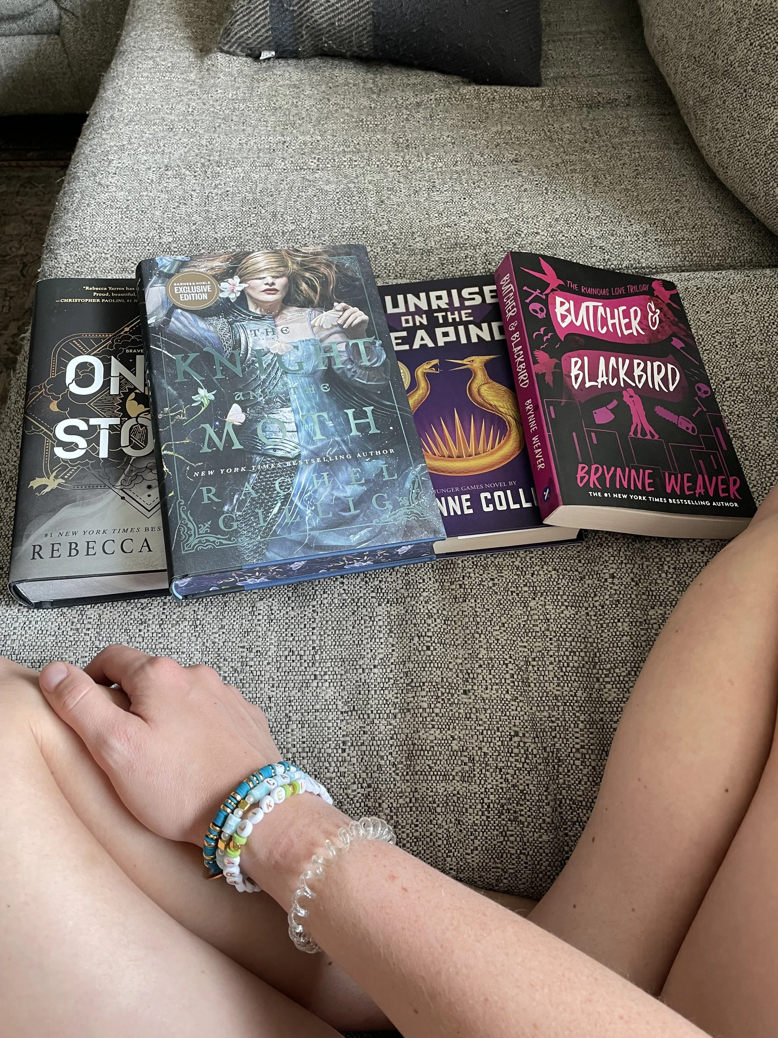 Four books with colorful covers resting on a gray fabric couch, with a person's arm and leg in the foreground, wearing beaded bracelets.