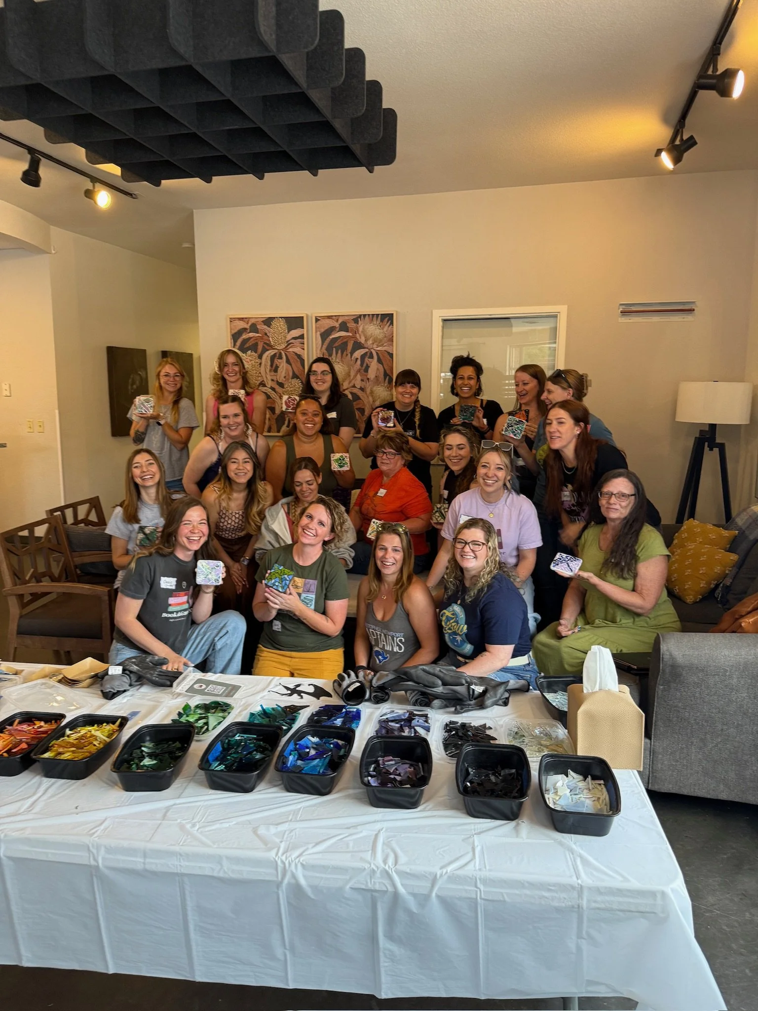 Group of women gathered around a table with art supplies, smiling and holding glass mosaic pieces, in a cozy indoor setting.