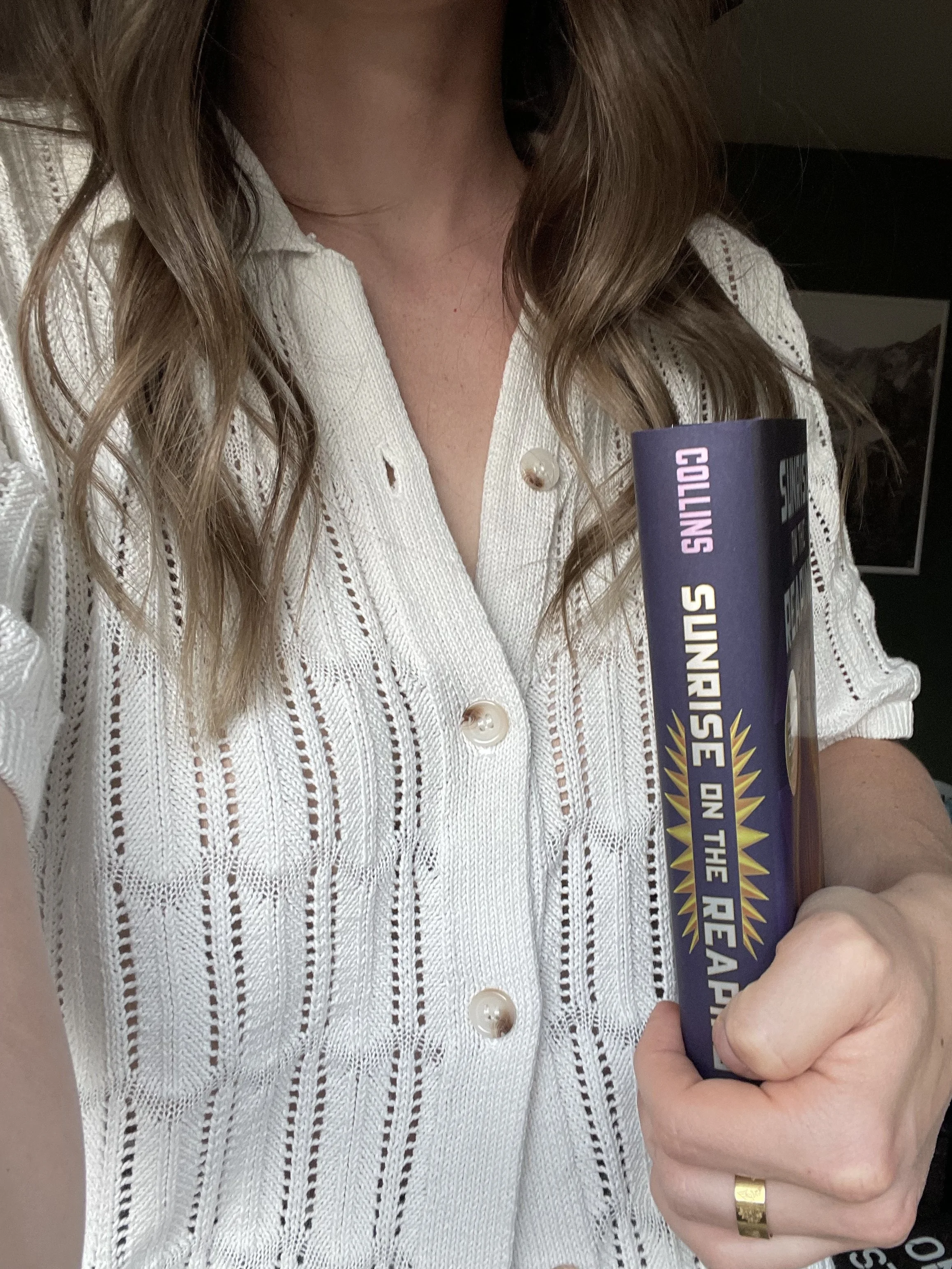 Person wearing a white lace button-up shirt holding a book titled "Sunrise on the Reaper".
