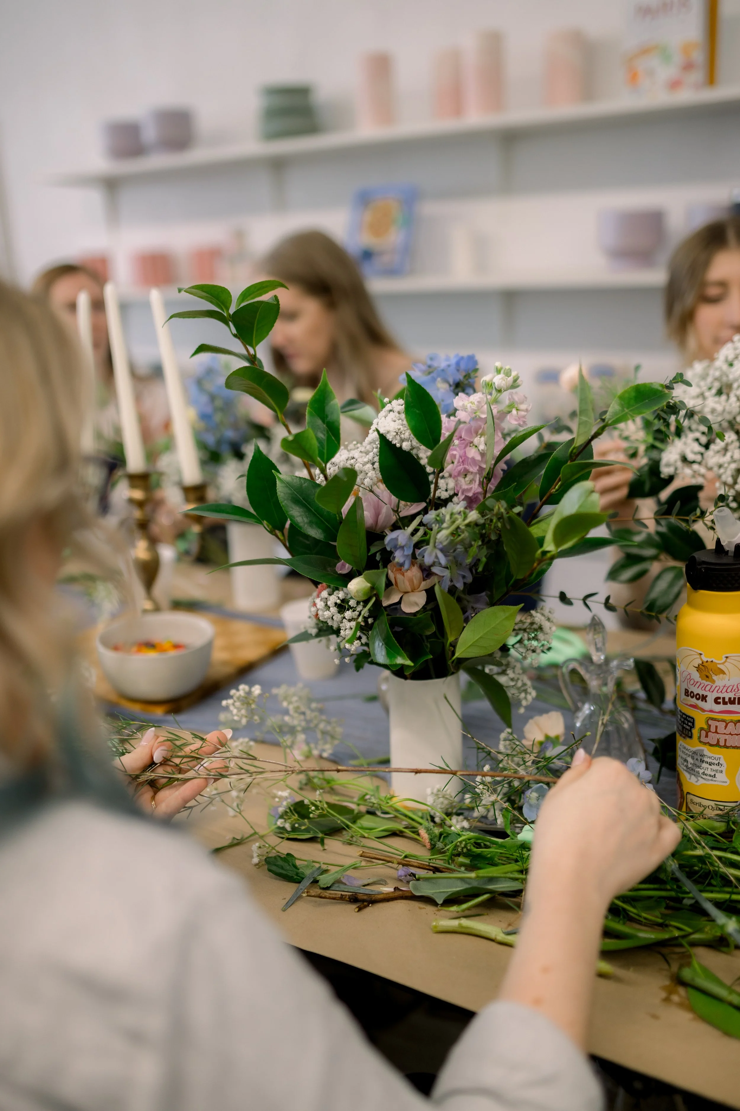 People creating floral arrangements at a table with various flowers and greenery in a cozy, decorated room.