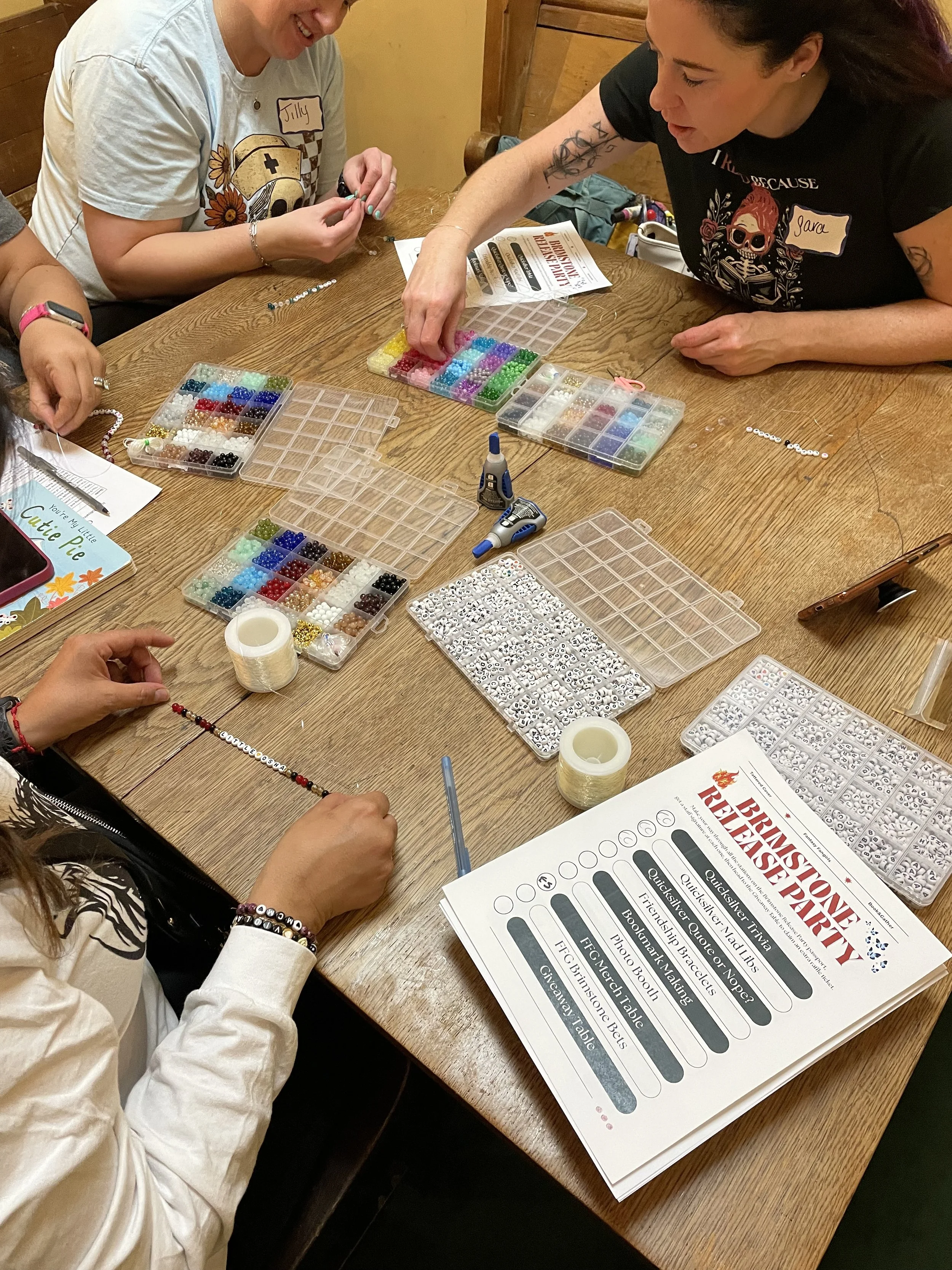 People making friendship bracelets with beads at a table at Tattered Cover Bookstore in Denver Colorado, during the Brimstone Relesae Party. There is a container of beads, a glue gun, and a printed guide for the activity.