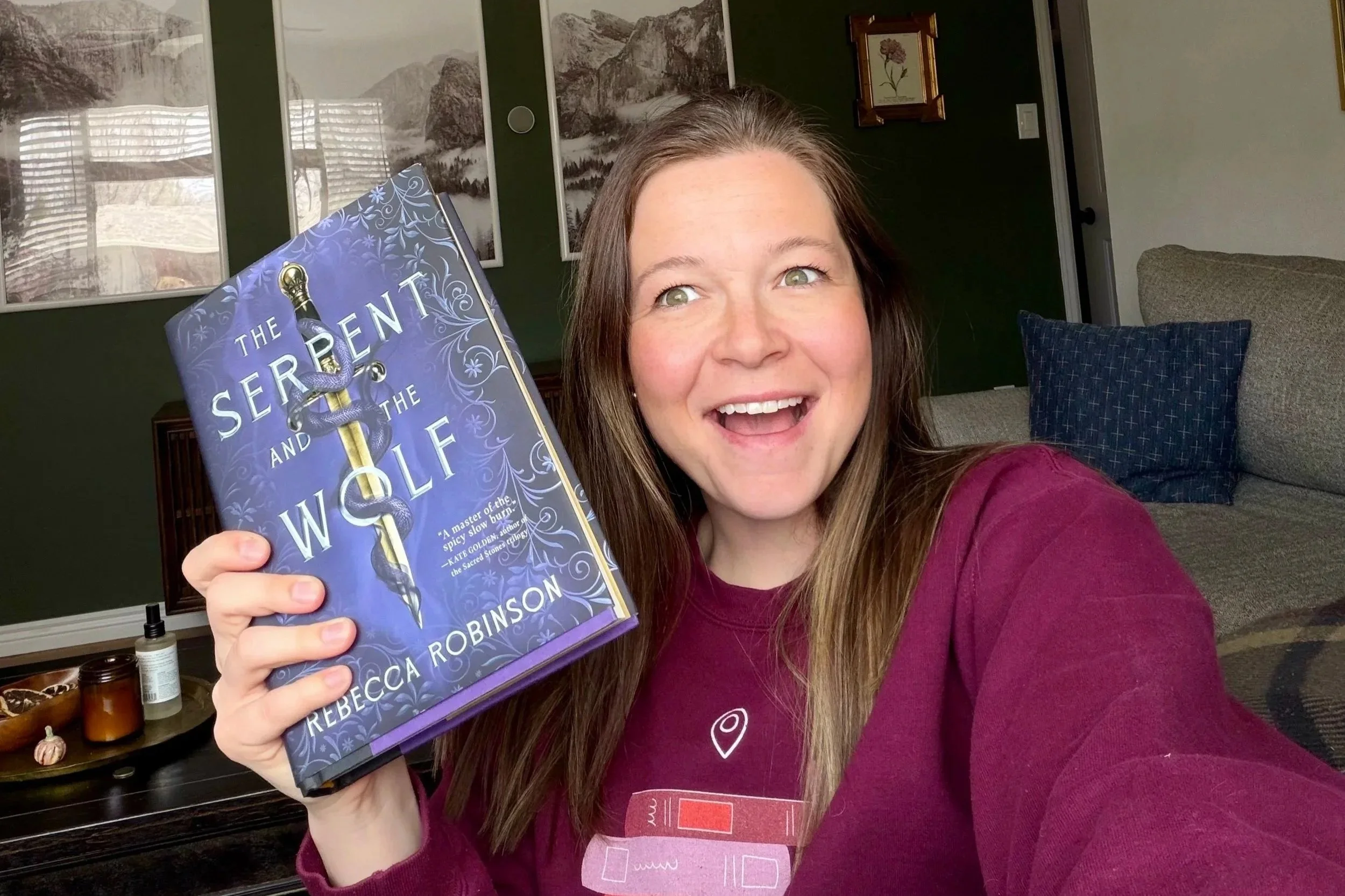 A woman with long brown hair and a pink sweater is holding a book titled 'The Serpent and the Wolf' by Rebecca Robinson, smiling widely in a cozy living room with framed pictures on the wall and a gray sofa with dark blue pillows in the background.