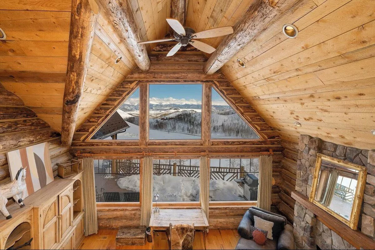 A cozy mountain cabin interior with a large triangular window showcasing snow-covered mountains and trees outside. The room features wooden walls, a ceiling fan, and a stone fireplace with a mirror above it. There is a small table with a chair and plush armchair inside.