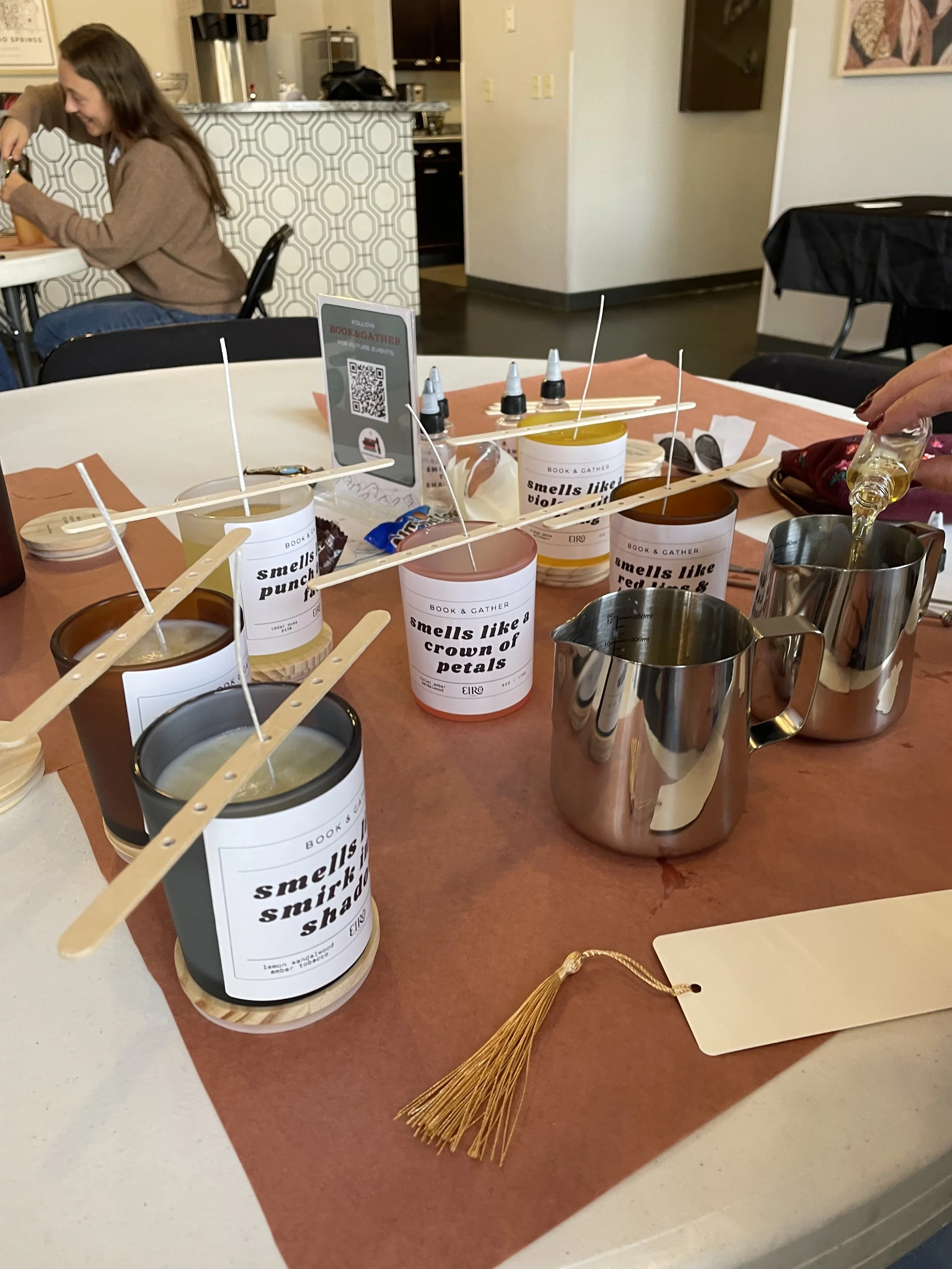 Table with scented candles labeled with various phrases, metal pitchers, and a gold tassel during Book&Gather's candle-making workshop.