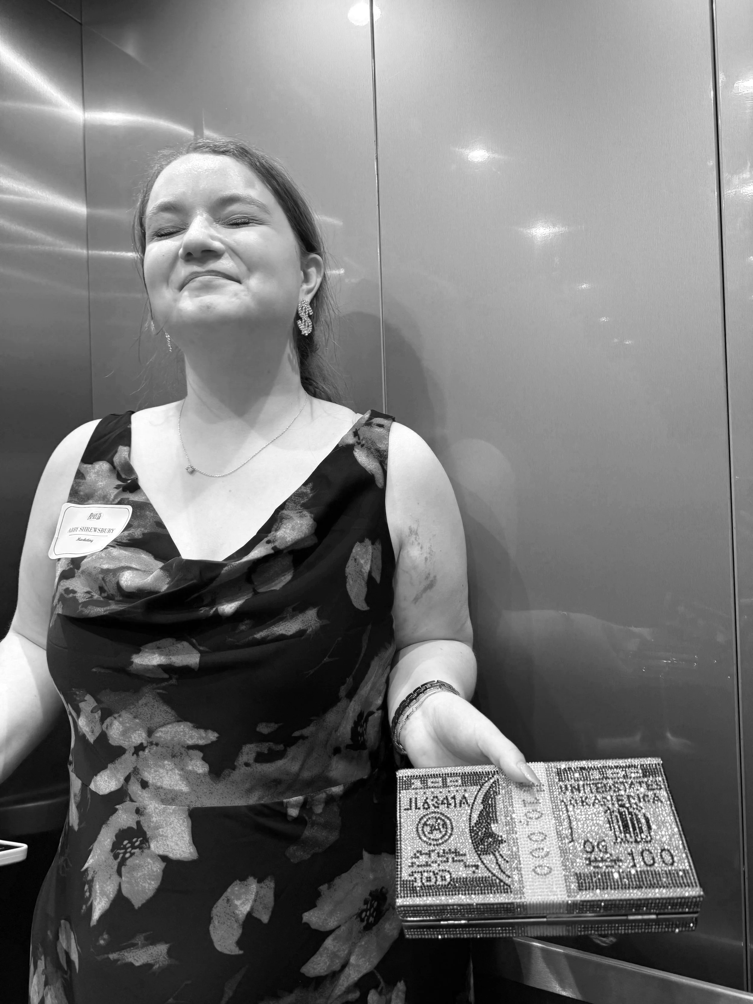A woman with a smile standing inside an elevator, holding a box designed to look like a dollar bill.
