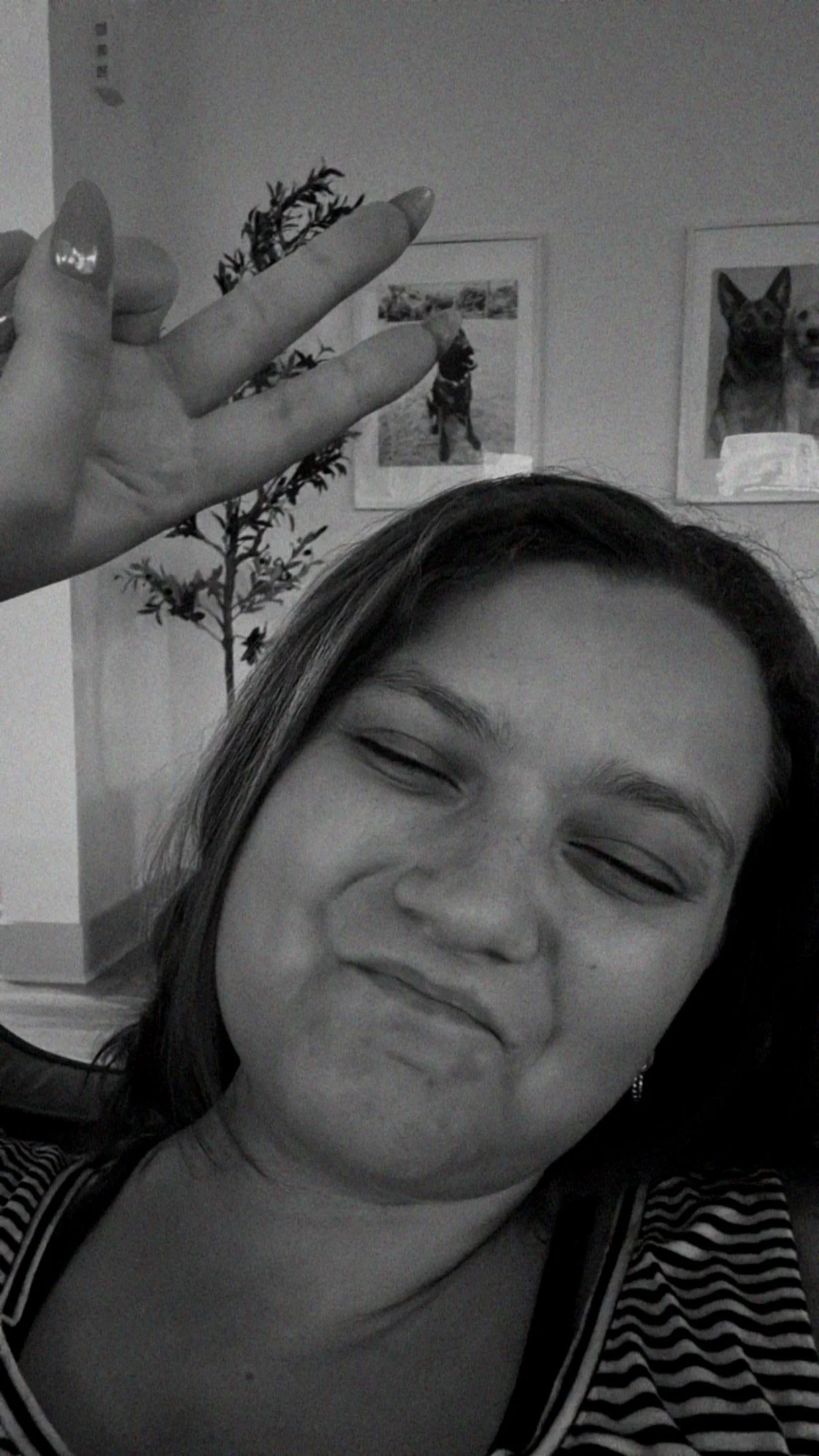 A woman making a scrunched face smile in a selfie, with her hand raised and fingers extended. In the background, framed pictures of dogs and a potted plant are visible.
