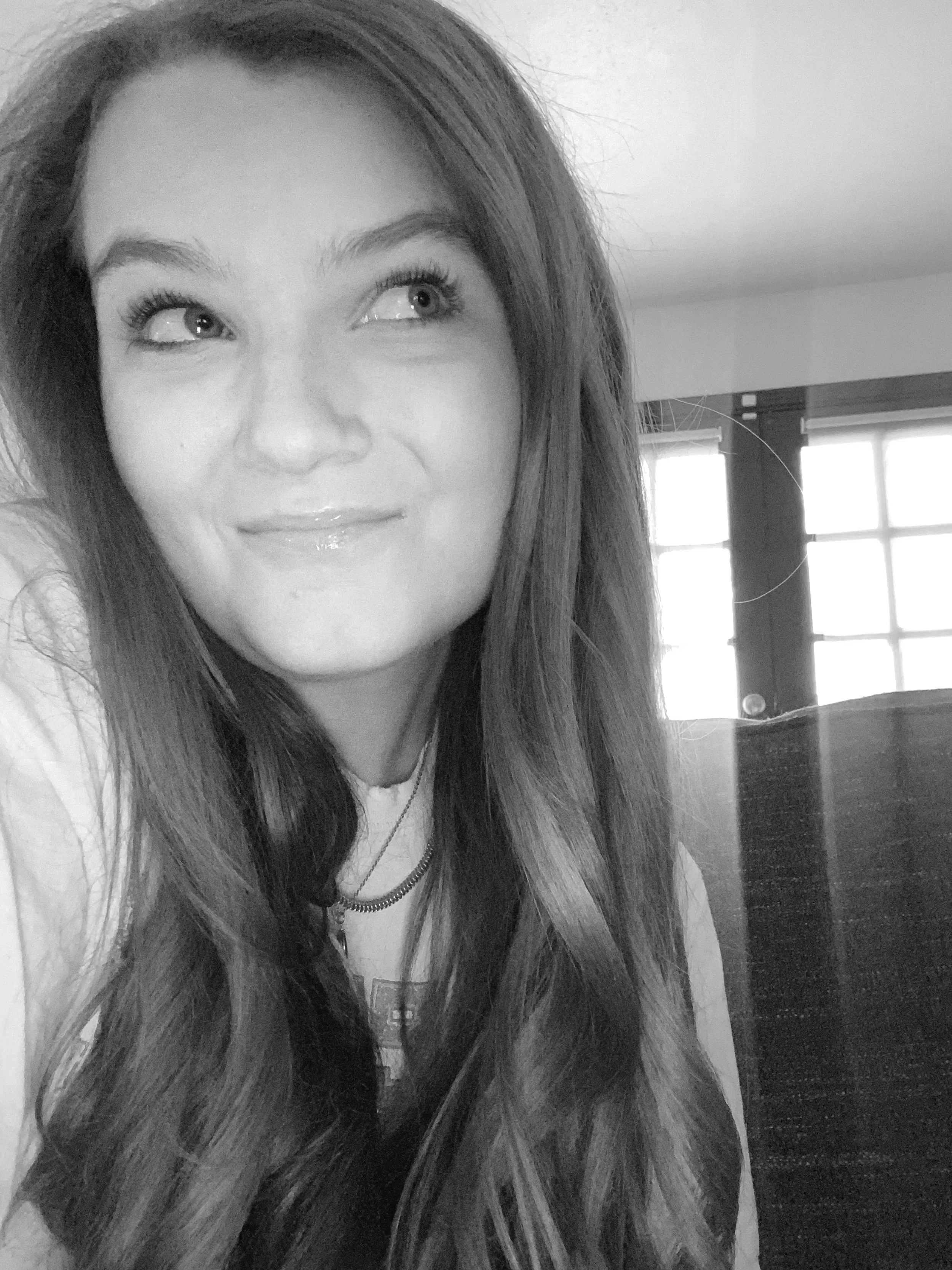 A young woman with long, wavy hair smiling and looking to the side, in black and white, indoors near a window.
