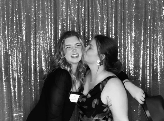 Two women smiling and hugging in front of a sparkly curtain backdrop at a photo booth.