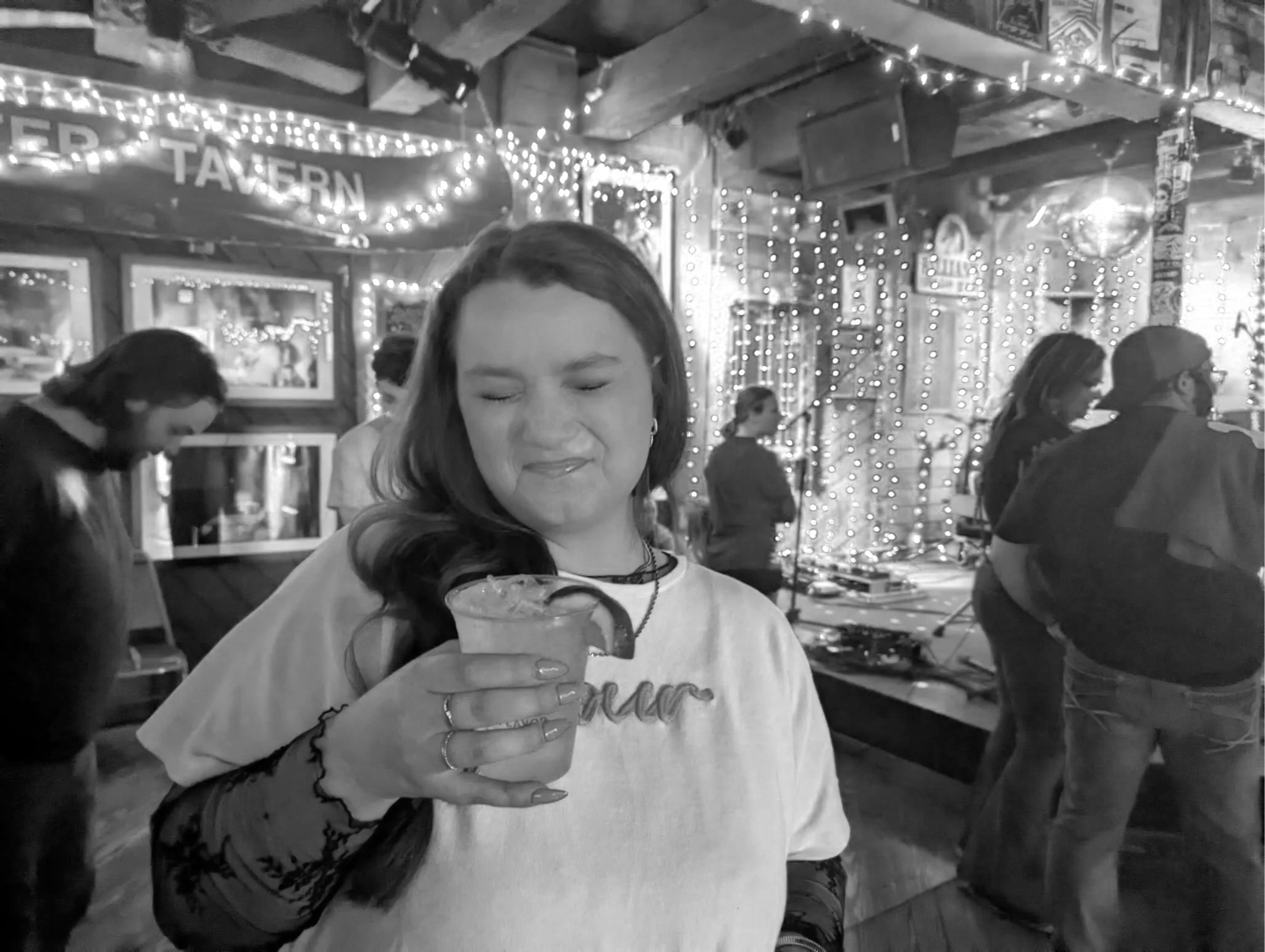 A woman holding a cocktail drink with a lemon wedge in a bar or restaurant with string lights and people in the background.