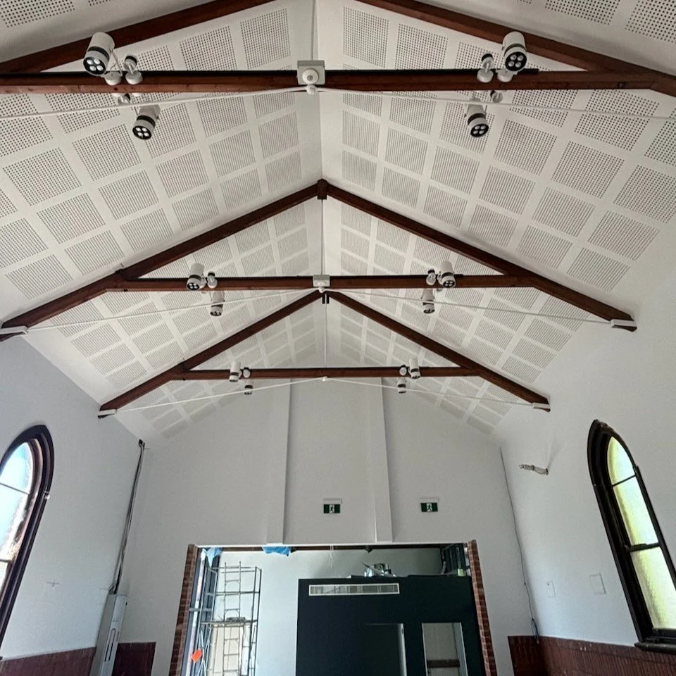 Ceiling in church with wooden beams and white acoustic panels, multiple spotlights and speakers, arched windows on side walls, a project completed by JAYQ Plaster & Trades.