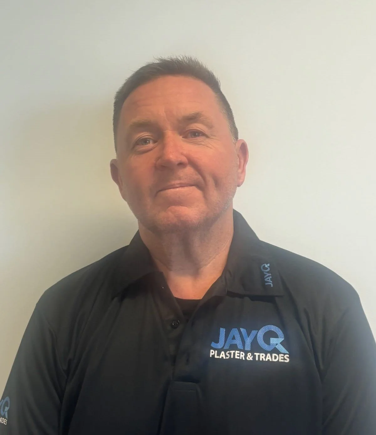 A man wearing a black polo shirt with the logo 'JAYQ PLASTER & TRADES' stands against a plain wall. Jason.