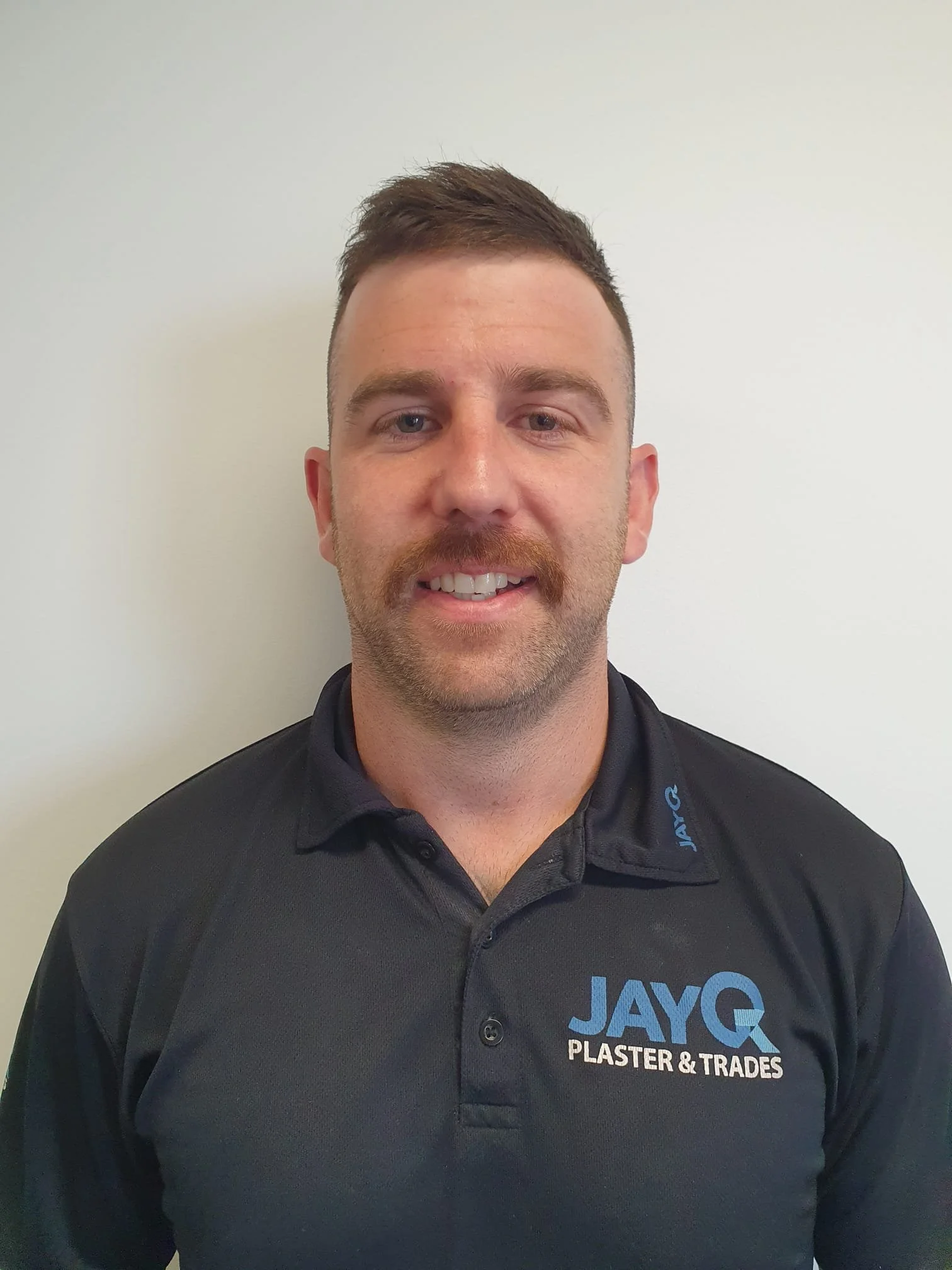 Headshot of a smiling man with short brown hair, a mustache, and a beard wearing a black polo shirt with the logo 'JAYQ PLASTER & TRADES' on it, standing against a plain light-colored wall. Steve.