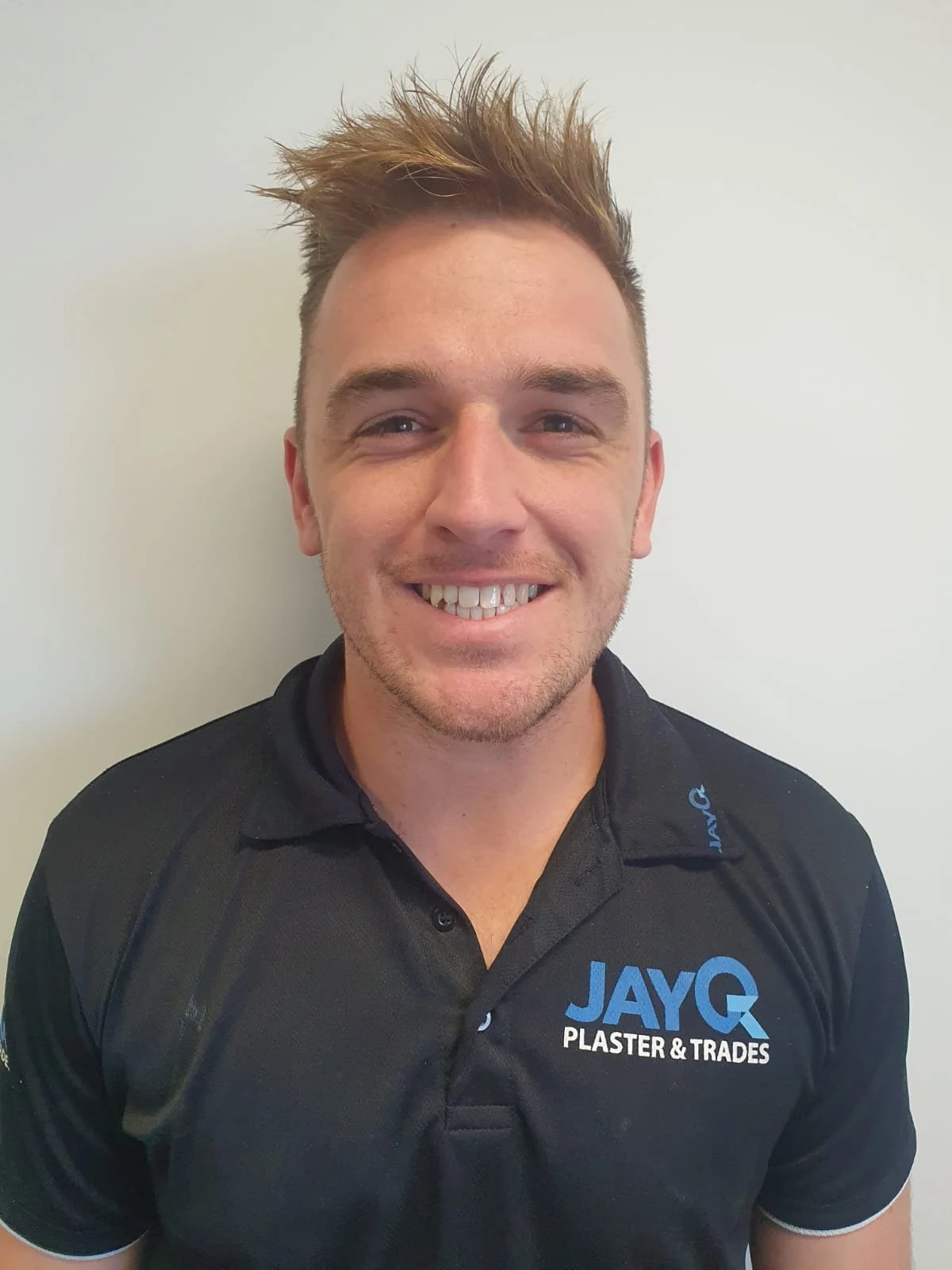 Smiling man in black polo shirt with JAYQ PLASTER & TRADES logo against plain white wall. Ryan.