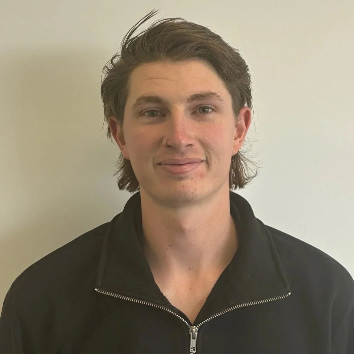 A young man with medium-length brown hair wearing a black zip-up jacket standing against a plain white wall. Luke from JAYQ Plaster & Trades.