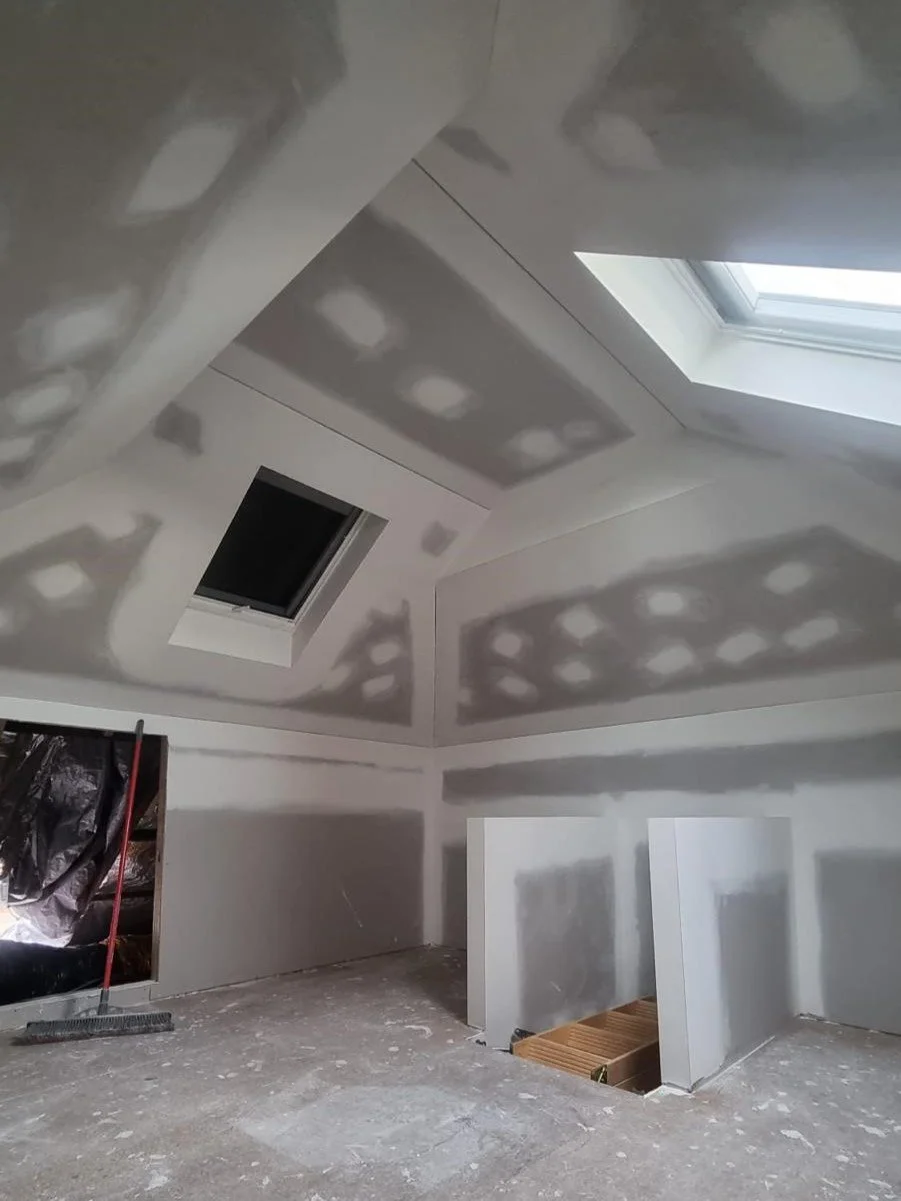An unfinished attic room with drywall installation, two skylights, and a small opening in the wall with construction materials and tools.