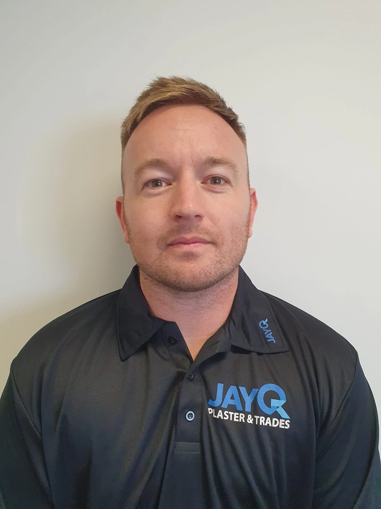 A man with short brown hair and a light beard wearing a black polo shirt with the logo 'JAYQ PLASTER & TRADES' on it, standing against a plain white wall. Dylan.