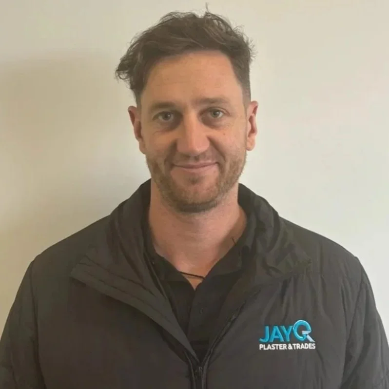A man with short, wavy hair and a light beard wearing a black jacket with a blue and white logo reading "JAYQ Plaster & Trades" standing against a plain white wall. Blade.